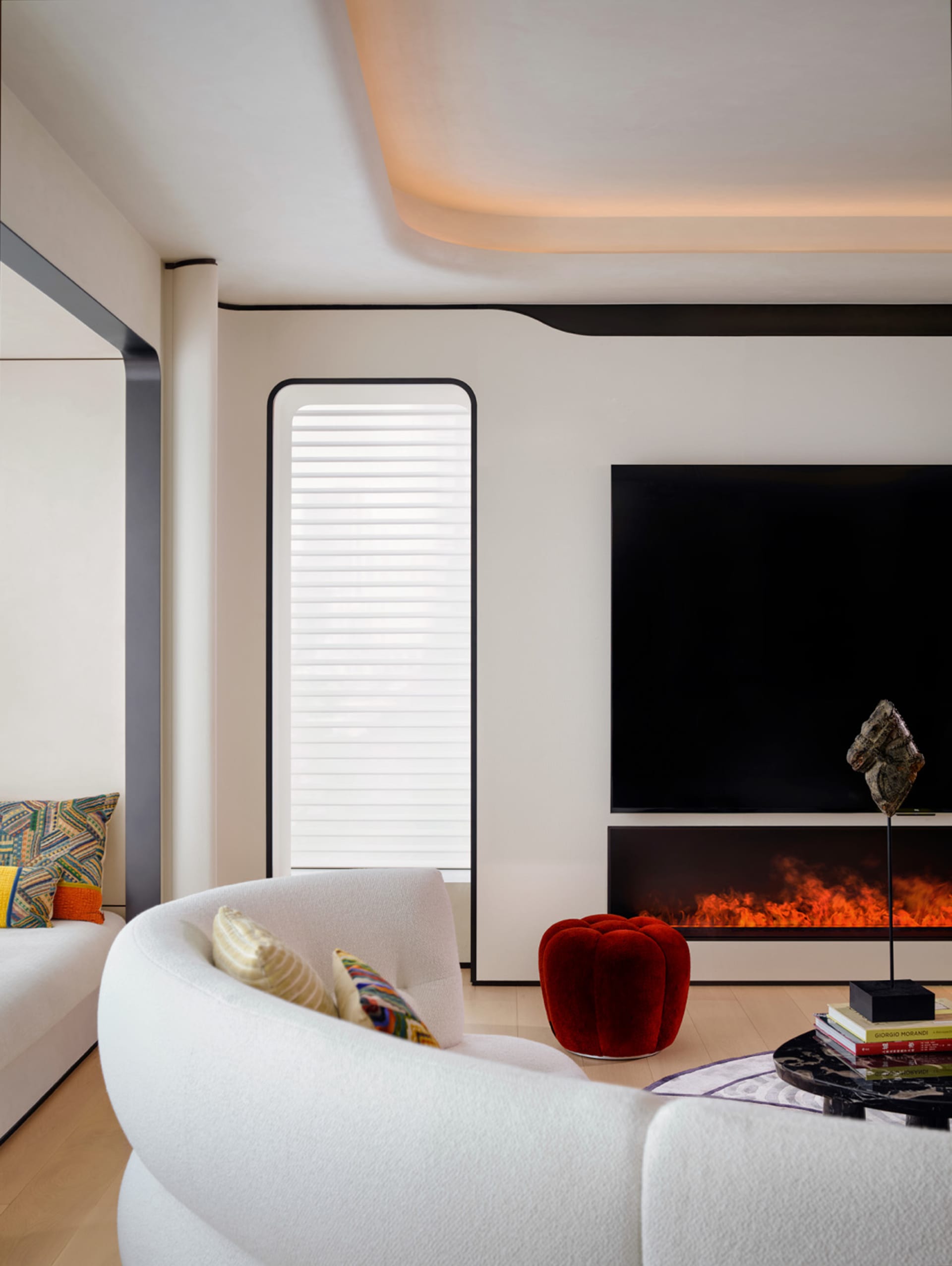 A modern living room featuring concrete walls and floor, with white curvilinear furniture, a red pouf, and a fireplace.