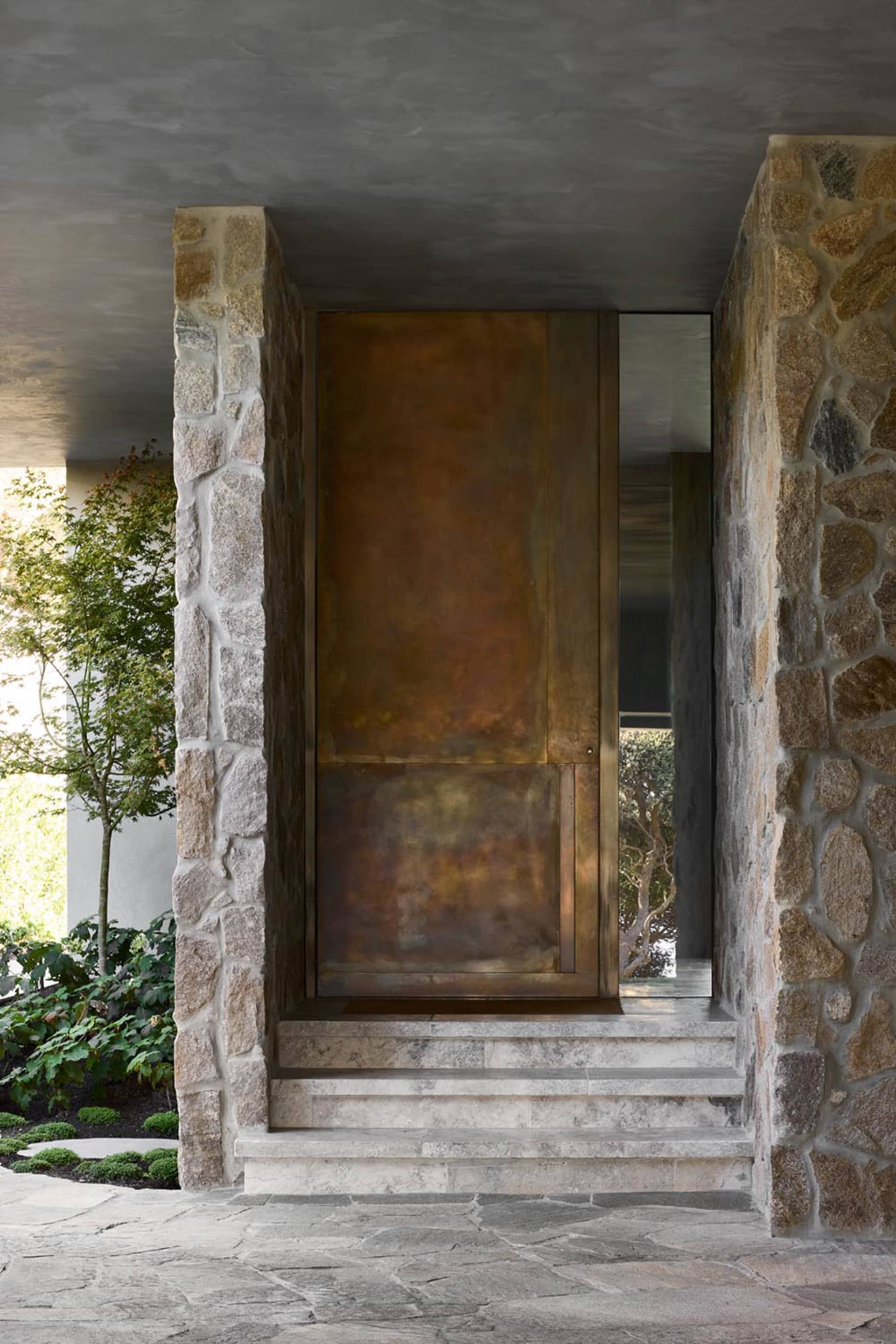 A modern stone door with a bronze finish, featuring a copper-colored metal panel, and surrounded by stone pillars and steps.