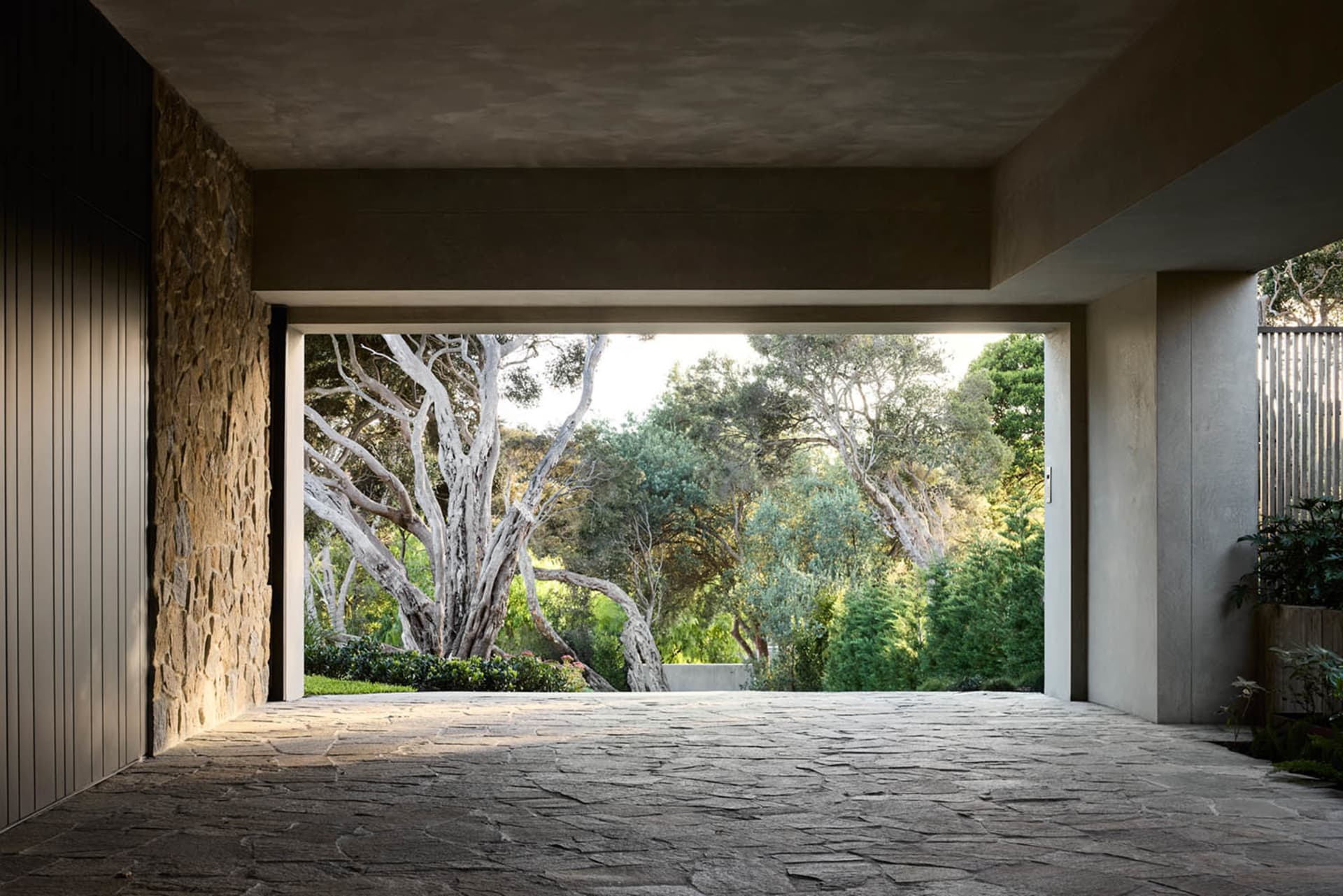 A modern mansion exterior with stone and concrete walls, featuring a large courtyard and surrounding trees.