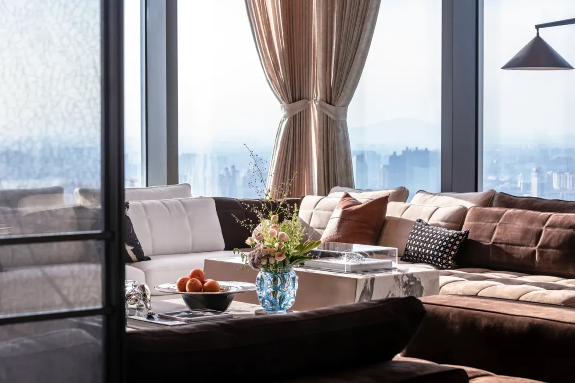 A modern living room featuring textured curtains, a large sectional sofa, and a coffee table with fruits and a vase of flower