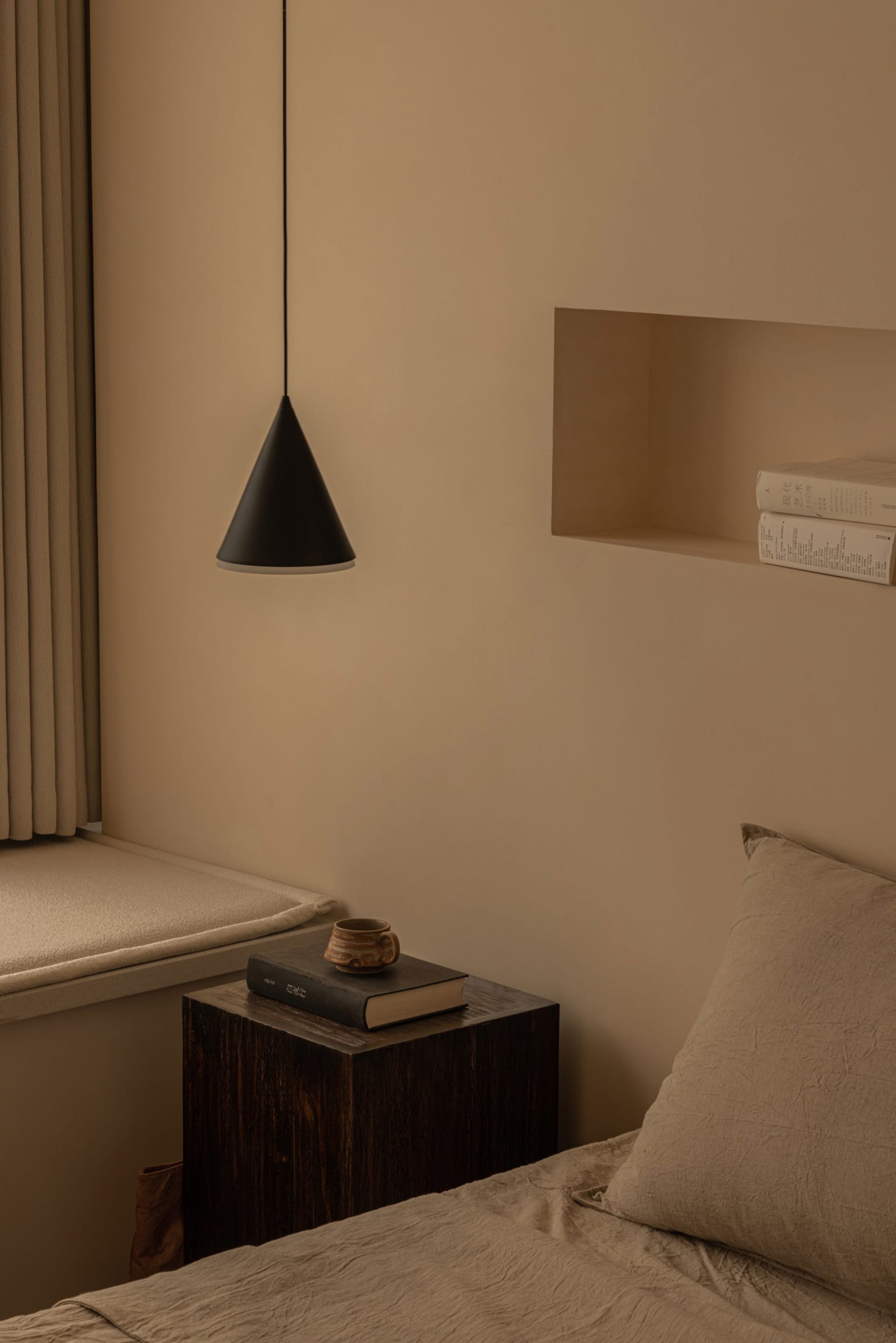 A minimalist bedroom featuring a wooden side table with a book and a ceramic mug, next to a window with beige curtains and a 