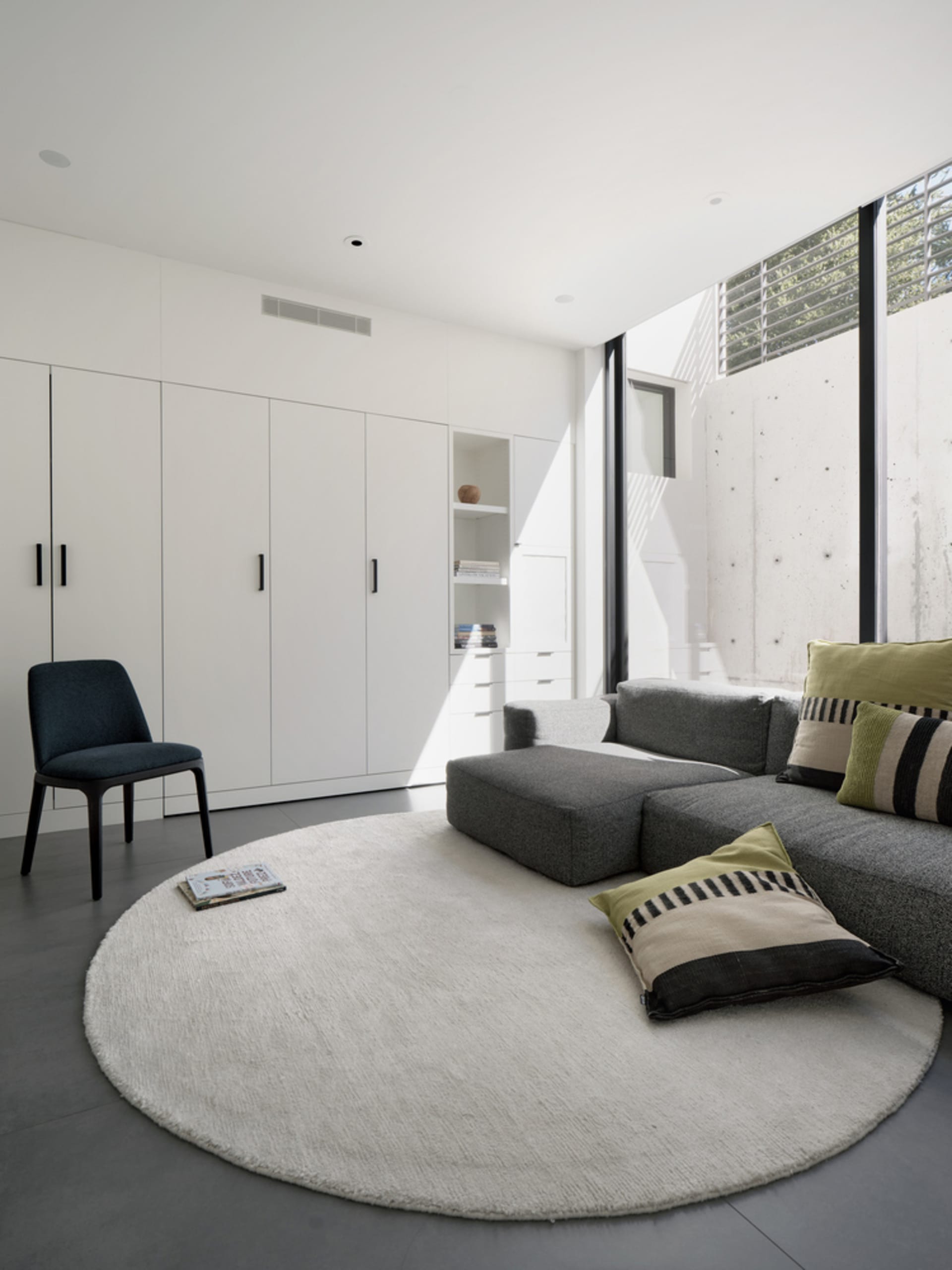 A minimalist living room with a circular white rug, a grey sofa with green and striped cushions, a grey ottoman, and a concre