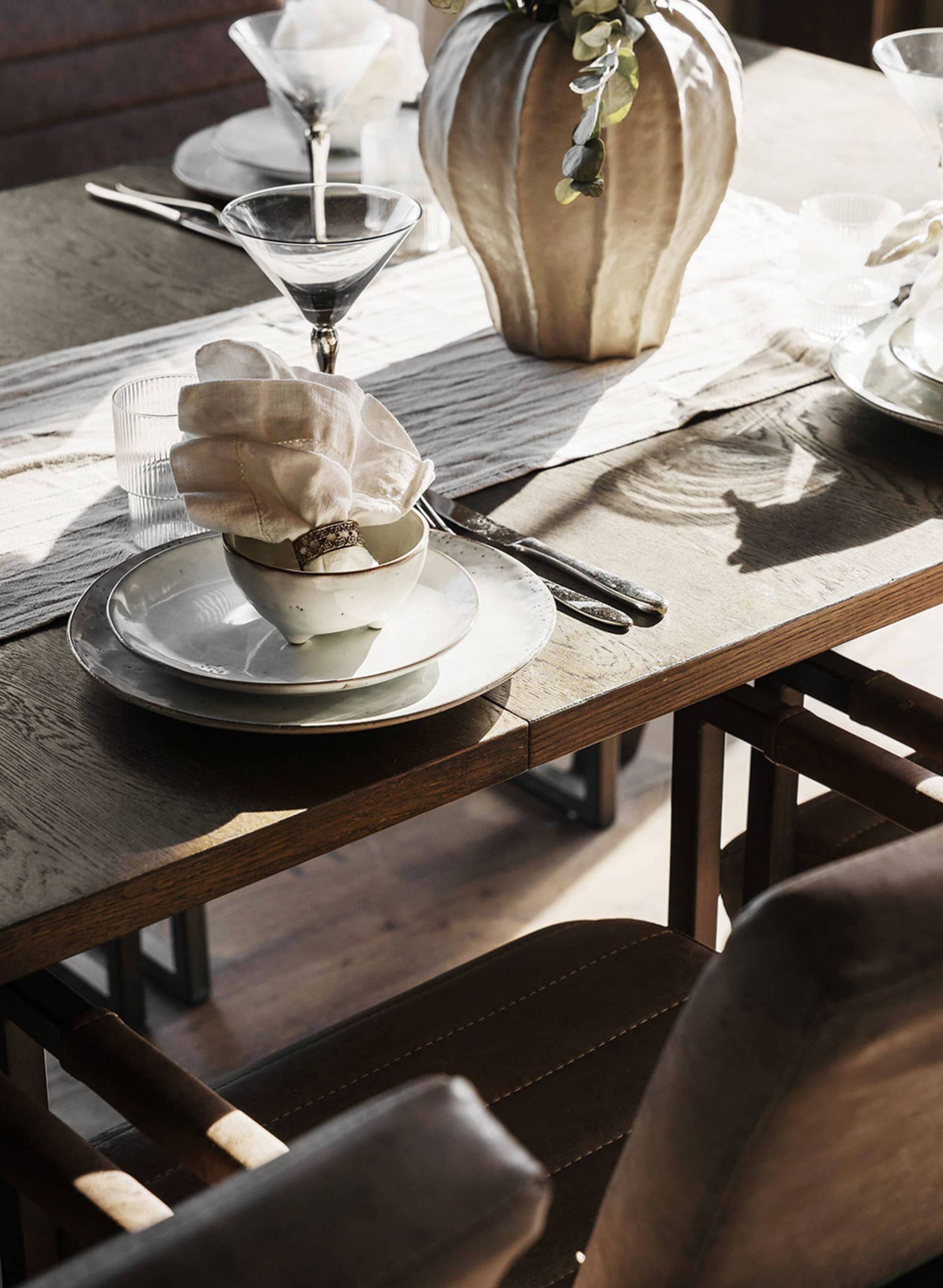 A modern dining room featuring wood furniture, white plates, and a gold ring on a napkin, with a vase and place settings.