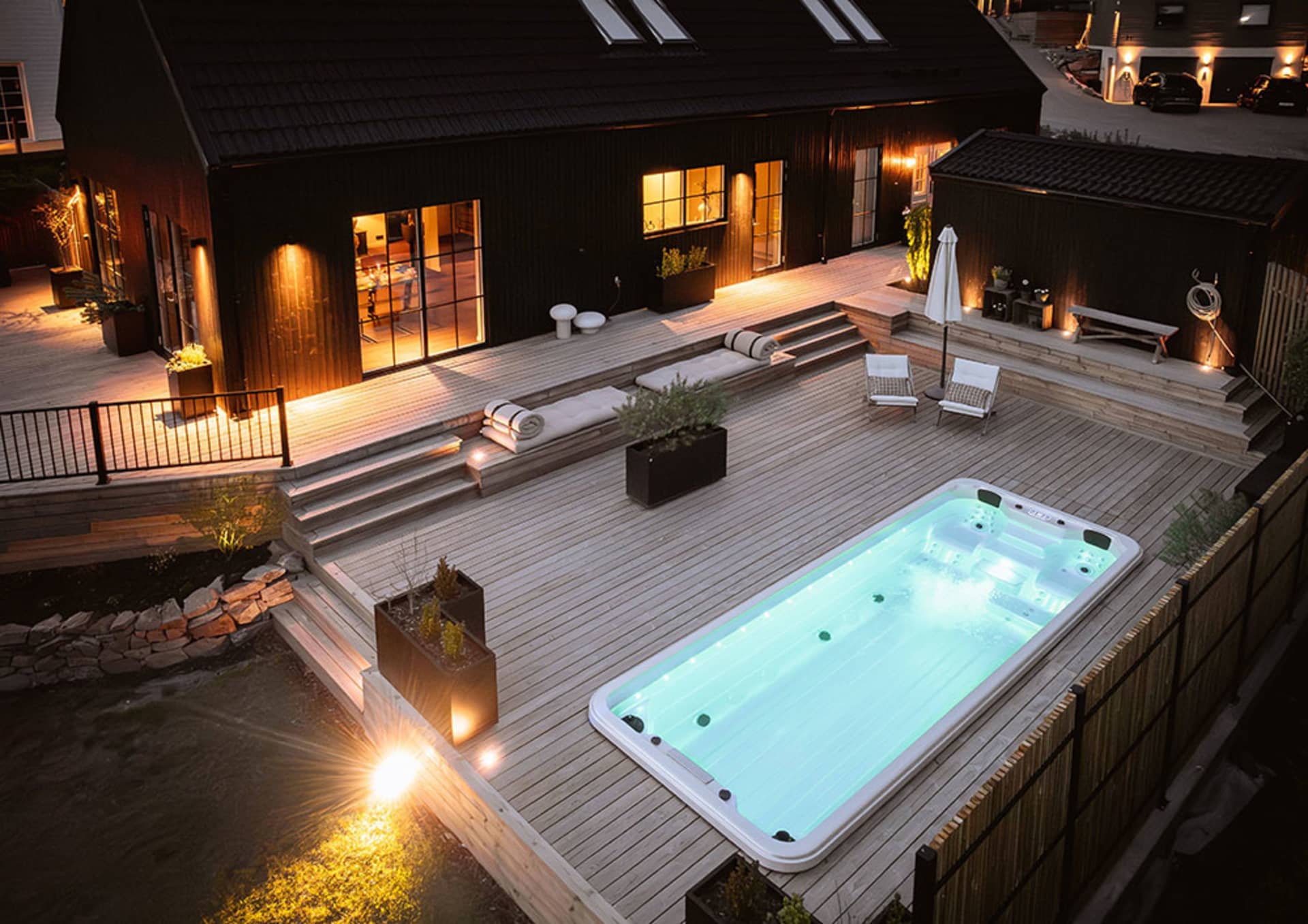 Modern residential backyard featuring a wooden terrace, concrete swimming pool, and stylish outdoor furniture, illuminated at