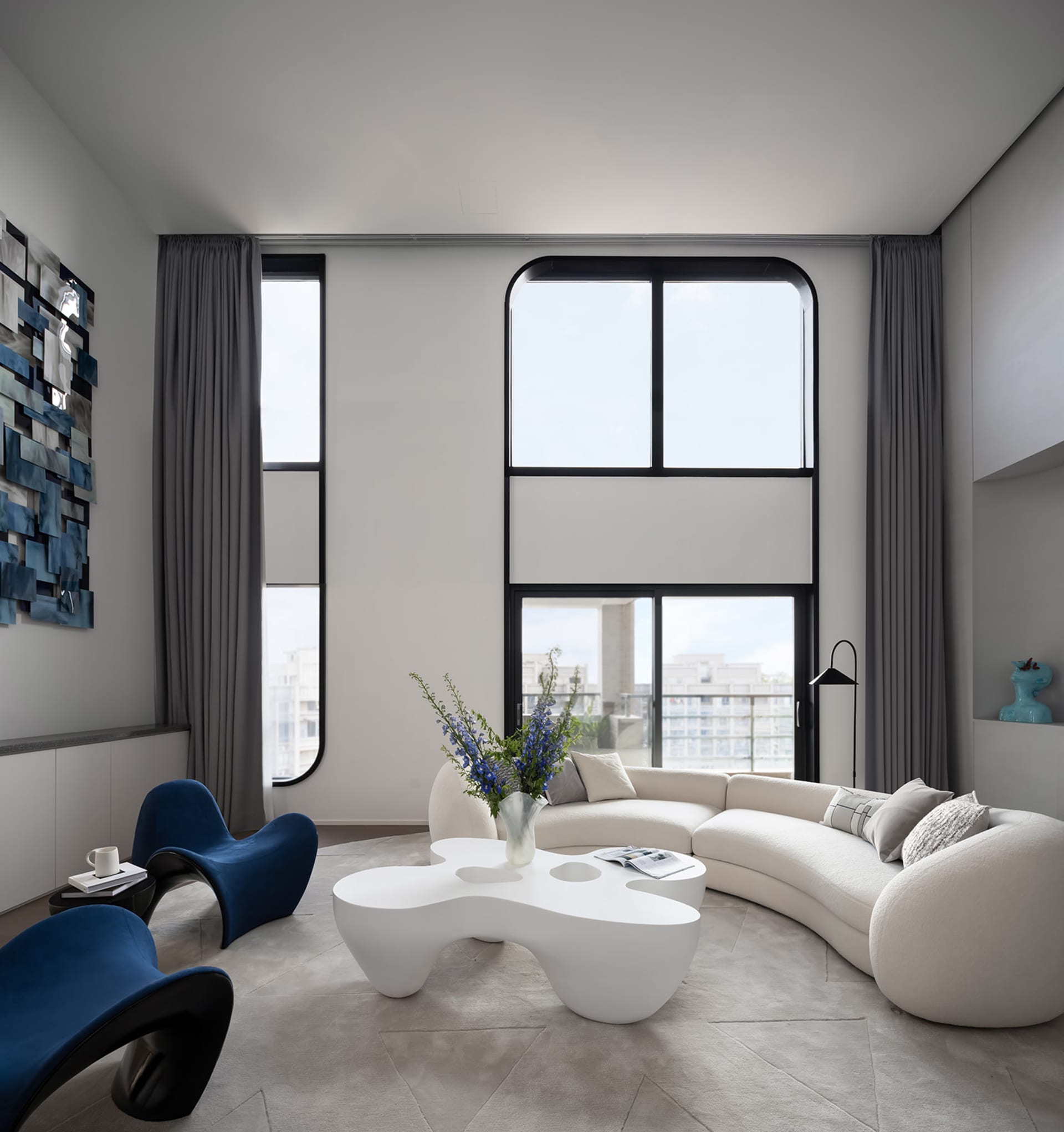 Modern living room with large windows featuring a white, curved sofa, blue armchairs, and a unique white coffee table with fl