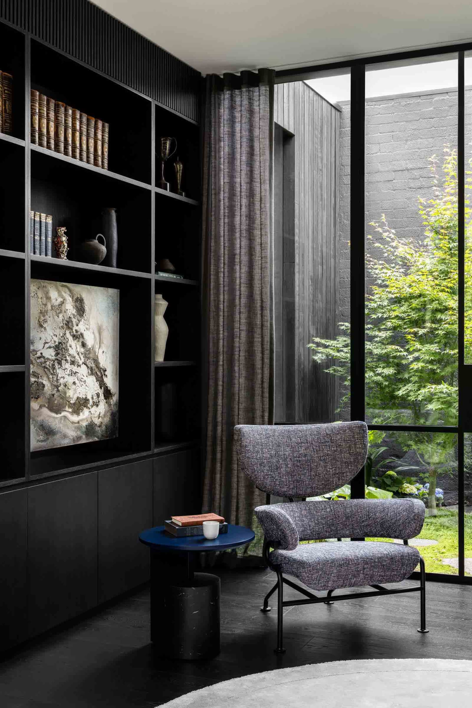 A modern library space featuring black wooden shelves, a cozy armchair, and a small circular table with a vase and a cup.