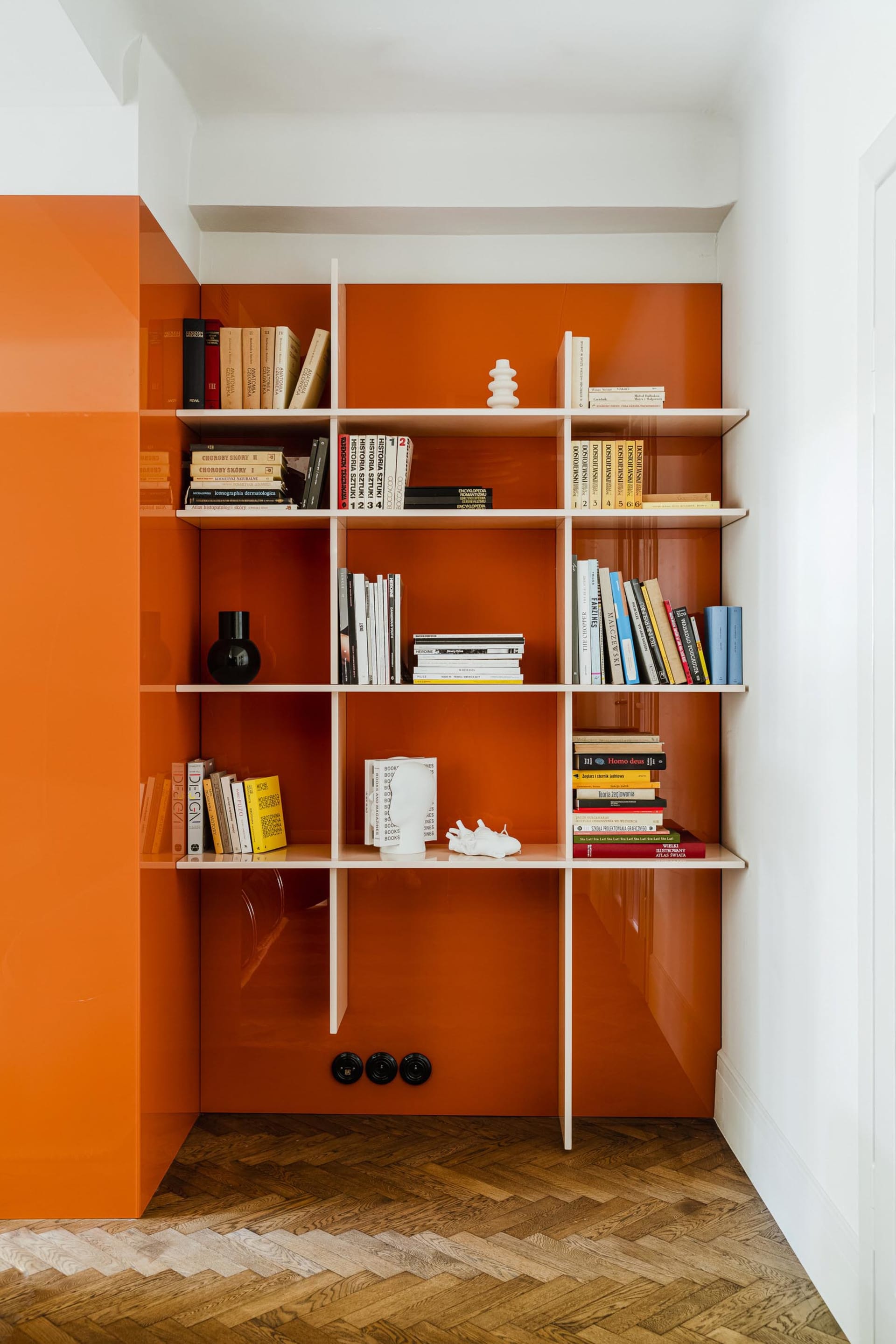 A modern, minimalist bookshelf with metallic orange panels and white shelves holding various books and items.