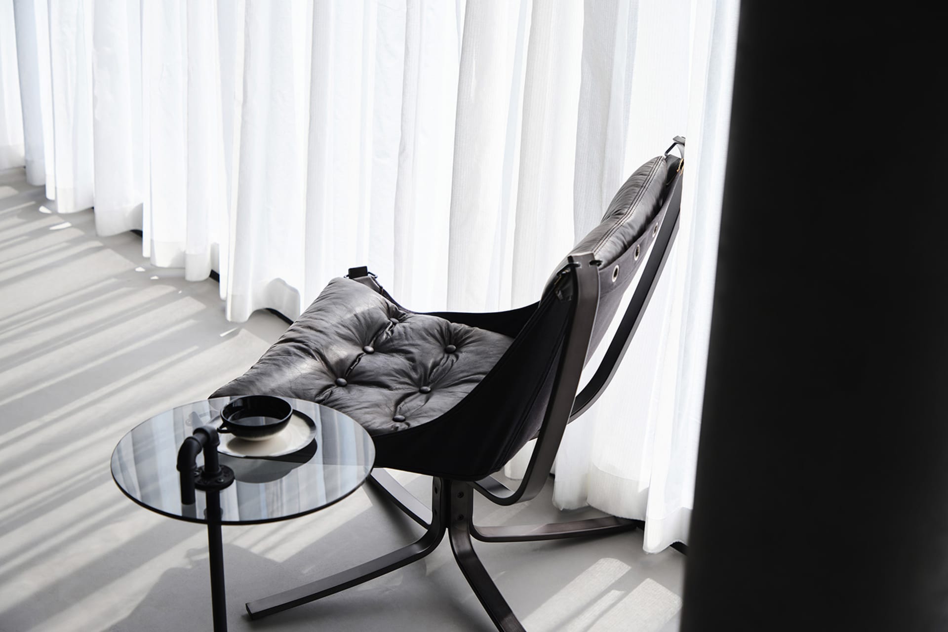 A sleek, modern leather chair with white cushions sits next to a round glass table with a mug and plate, against a backdrop o