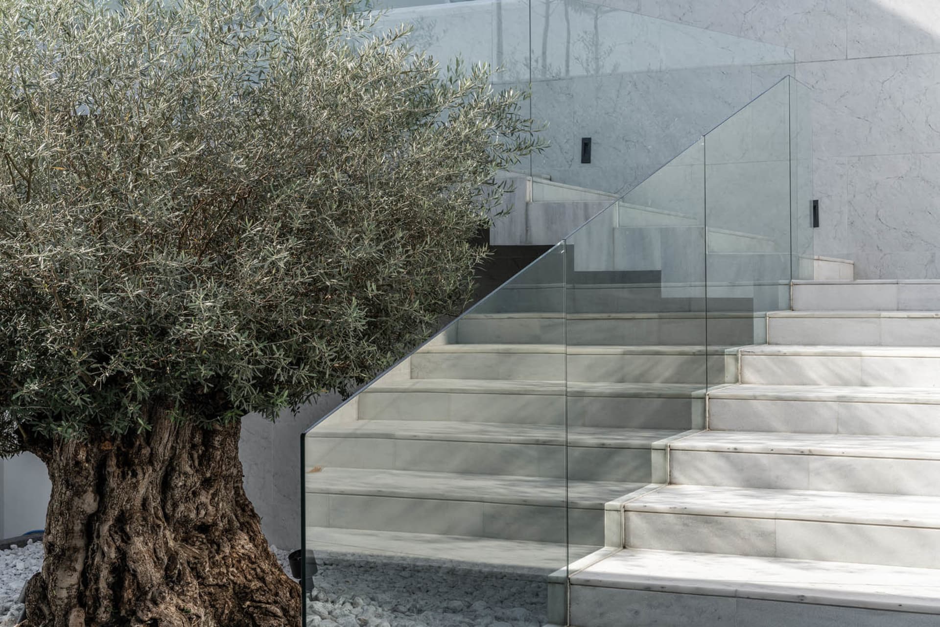 A modern staircase with marble steps and a glass railing, set against a concrete base, surrounded by greenery.