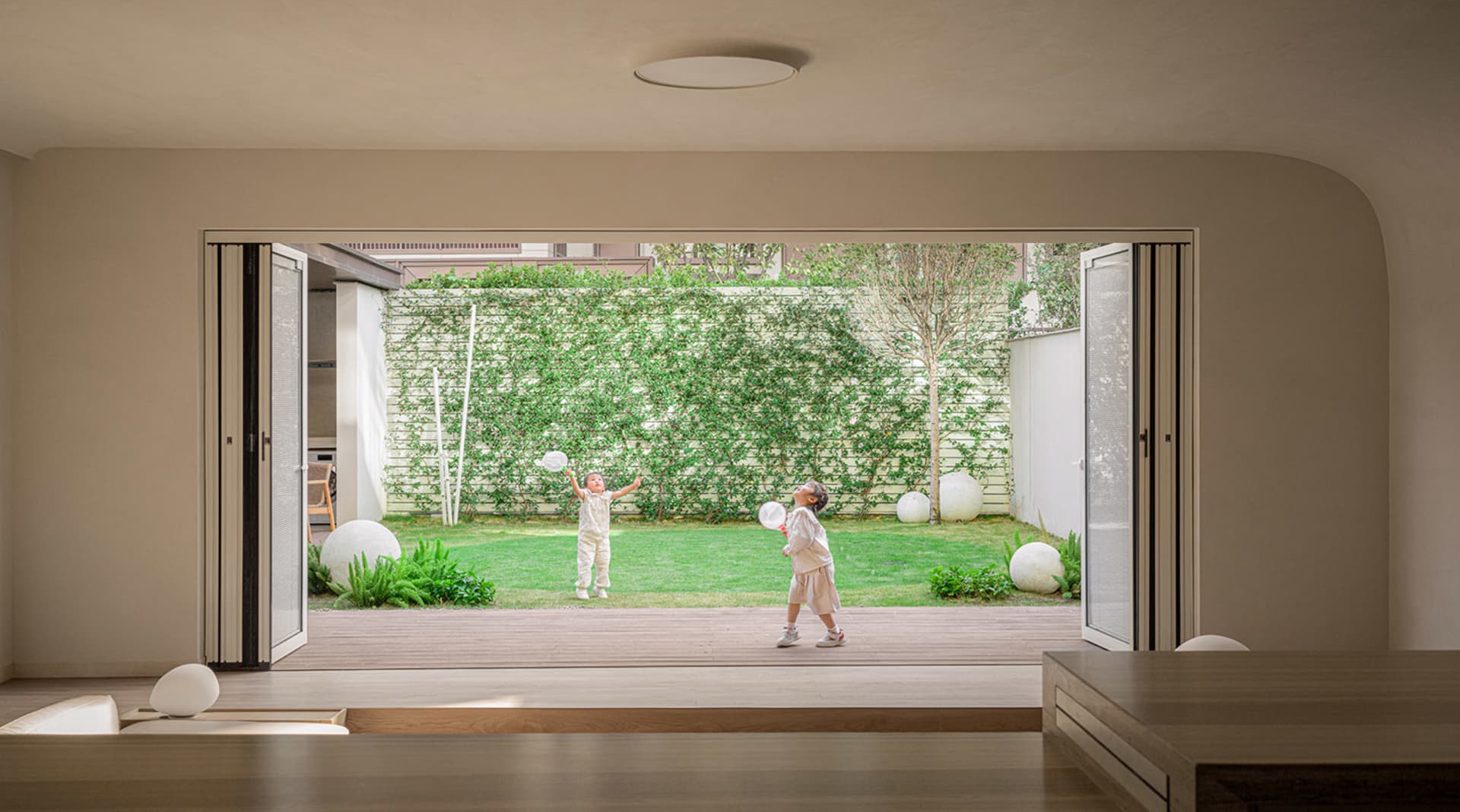 Two children playing badminton outdoors in a modern residential playarea with wooden deck and greenery.