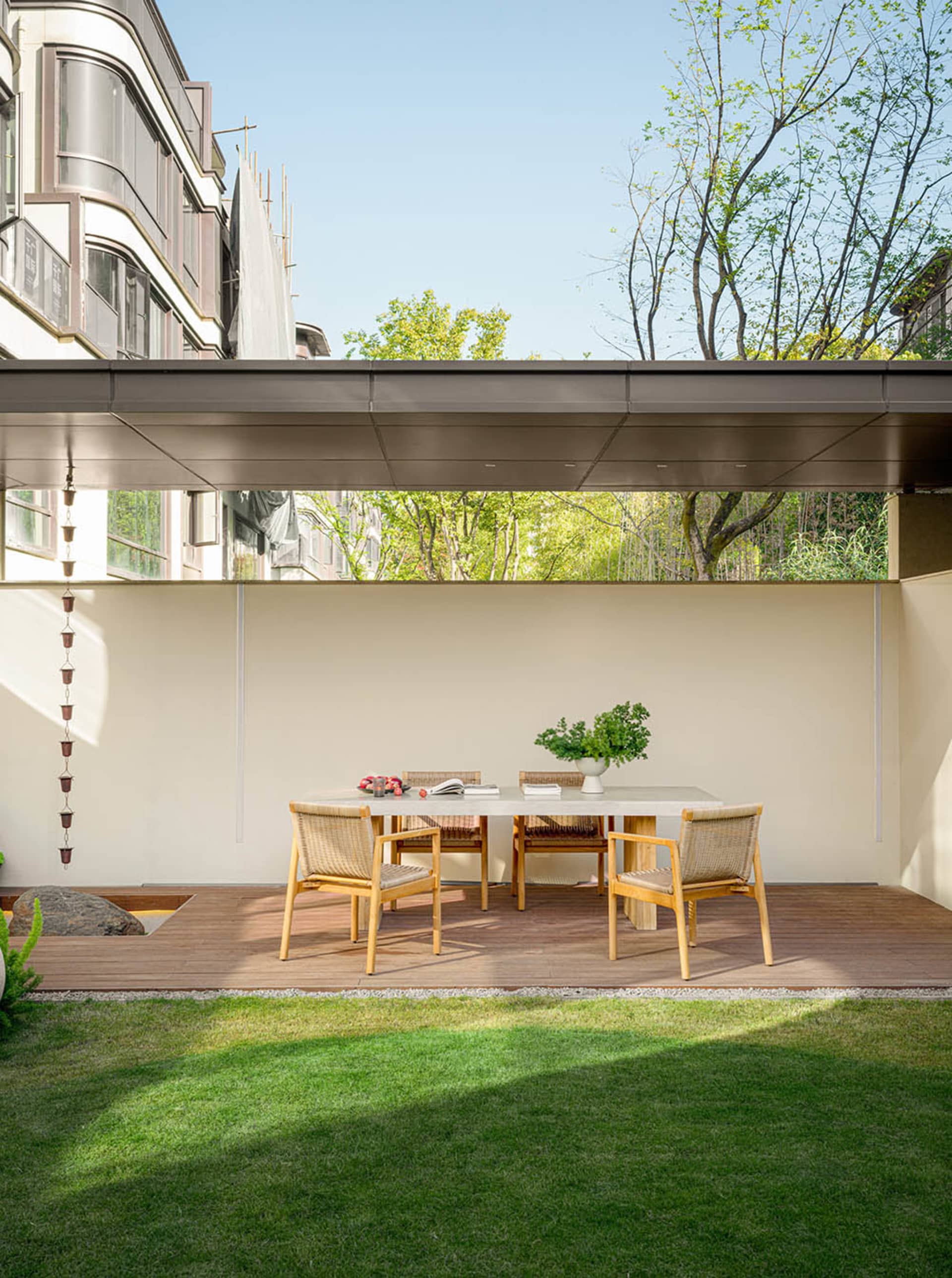 Modern terrace featuring a wooden table with a vase of flowers, surrounded by greenery and a wooden deck, with a gravel pathw