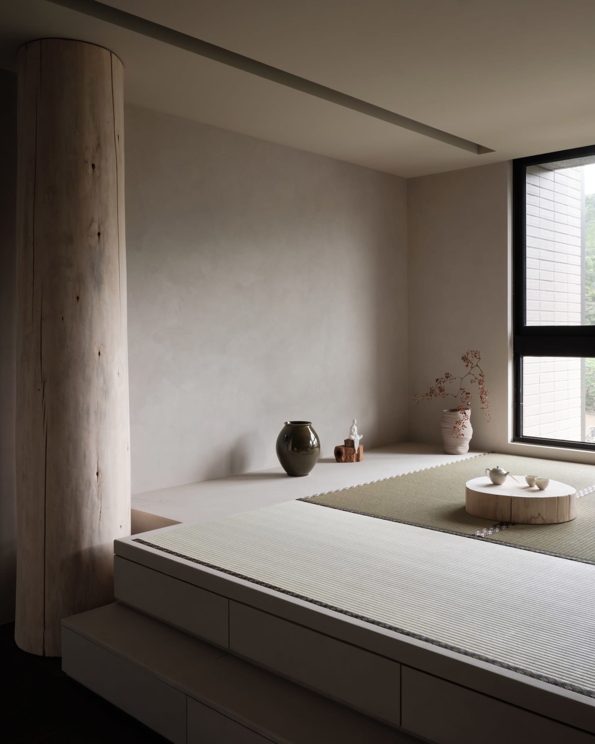 A minimalist living room featuring wooden furniture, a tatami mat, and a small tea set on a wooden platform.