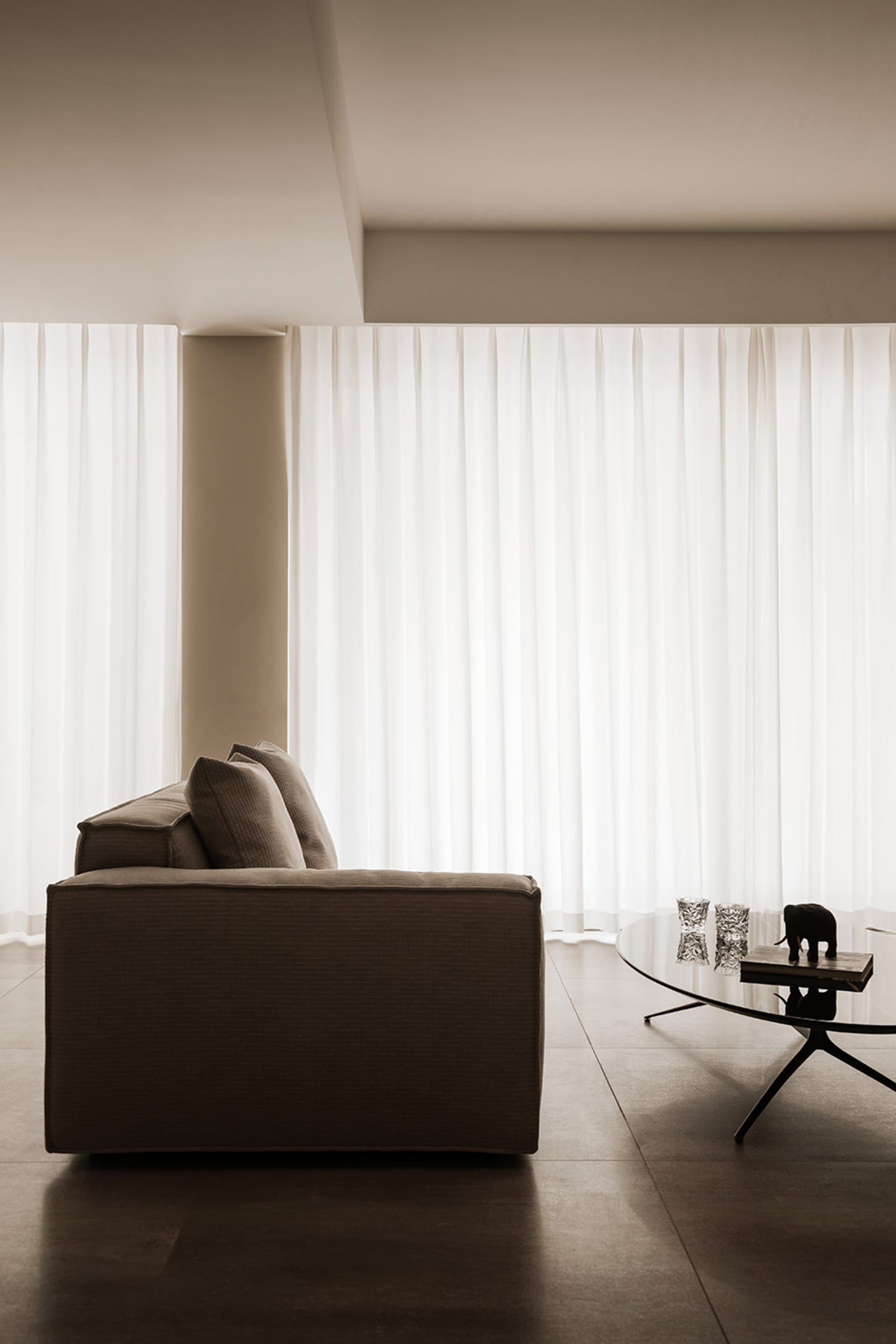 A minimalist living room with a brown upholstered sofa and white sheer curtains on the window, creating a serene and modern a