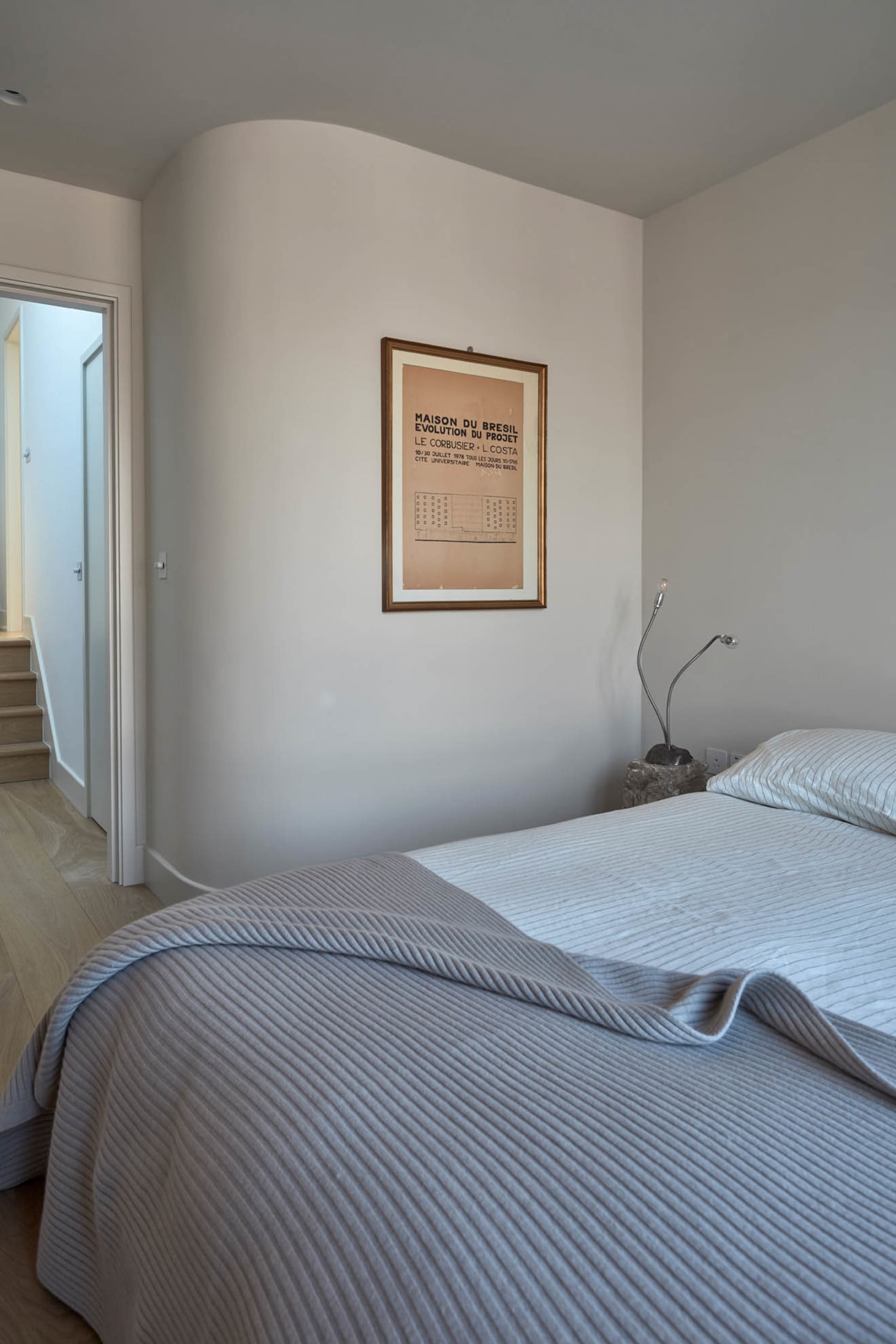 A minimalist bedroom with a light gray blanket on the bed, framed artwork on the wall, and modern lighting.