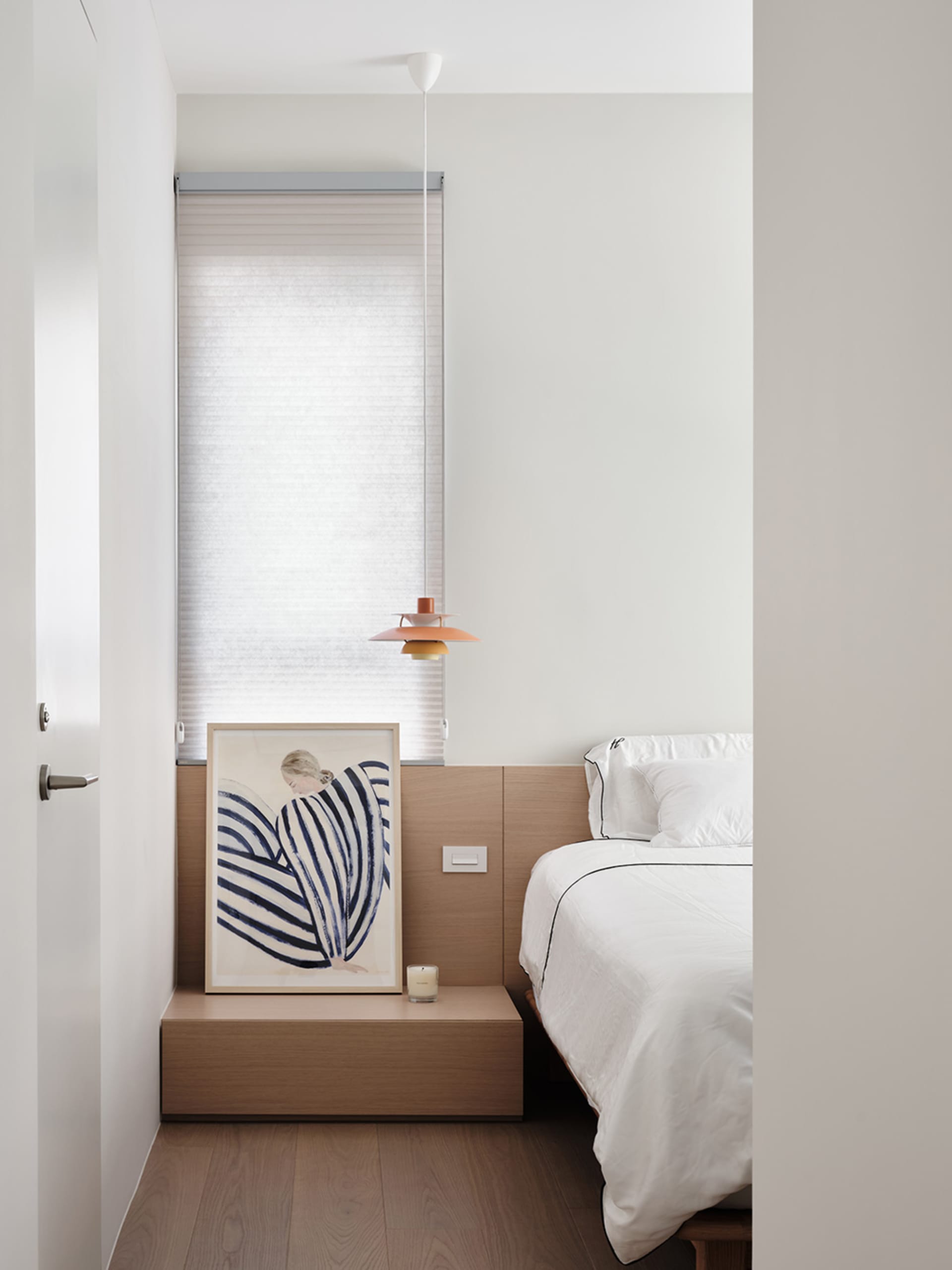 A minimalist bedroom with a wooden headboard, a white bed, and a framed abstract painting.