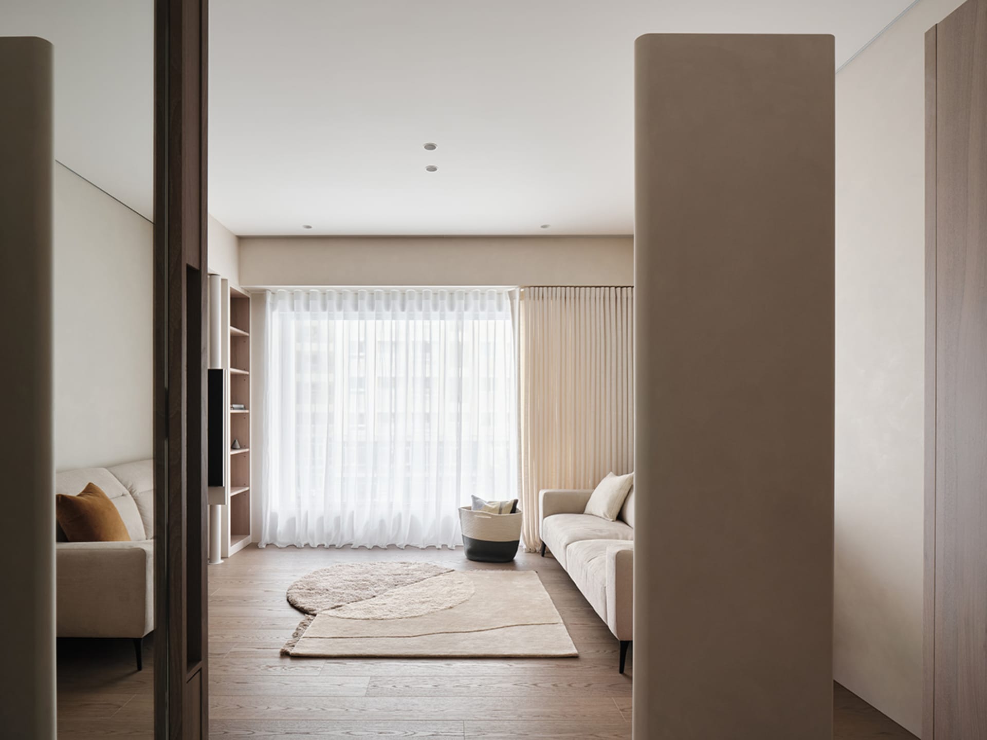A modern living room featuring neutral-colored furniture, a patterned rug, and large windows with sheer curtains.