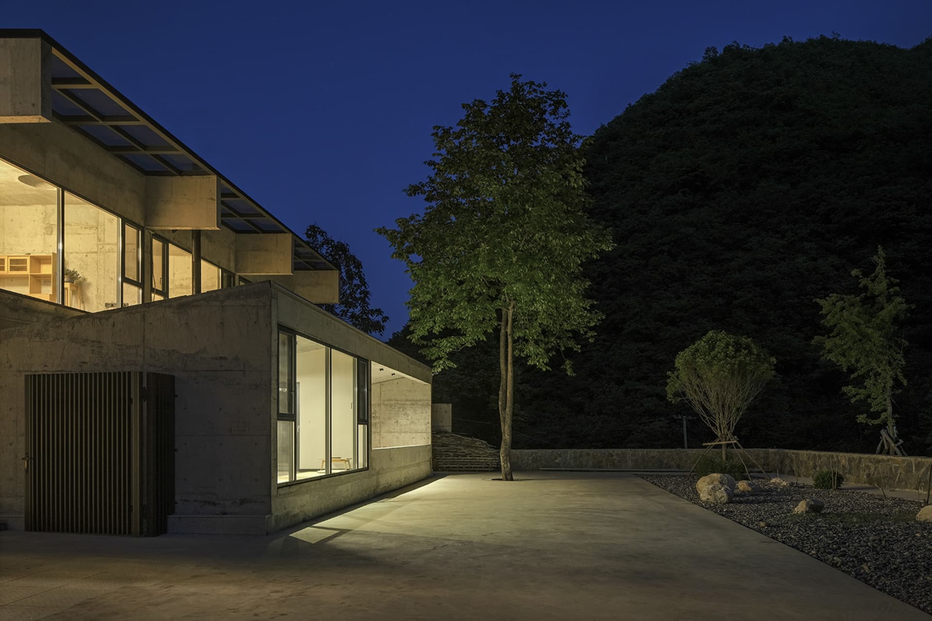 夜間照明下的住宅建築,擁有混凝土牆面與玻璃窗戶,周圍環繞花園、樹木與岩石。