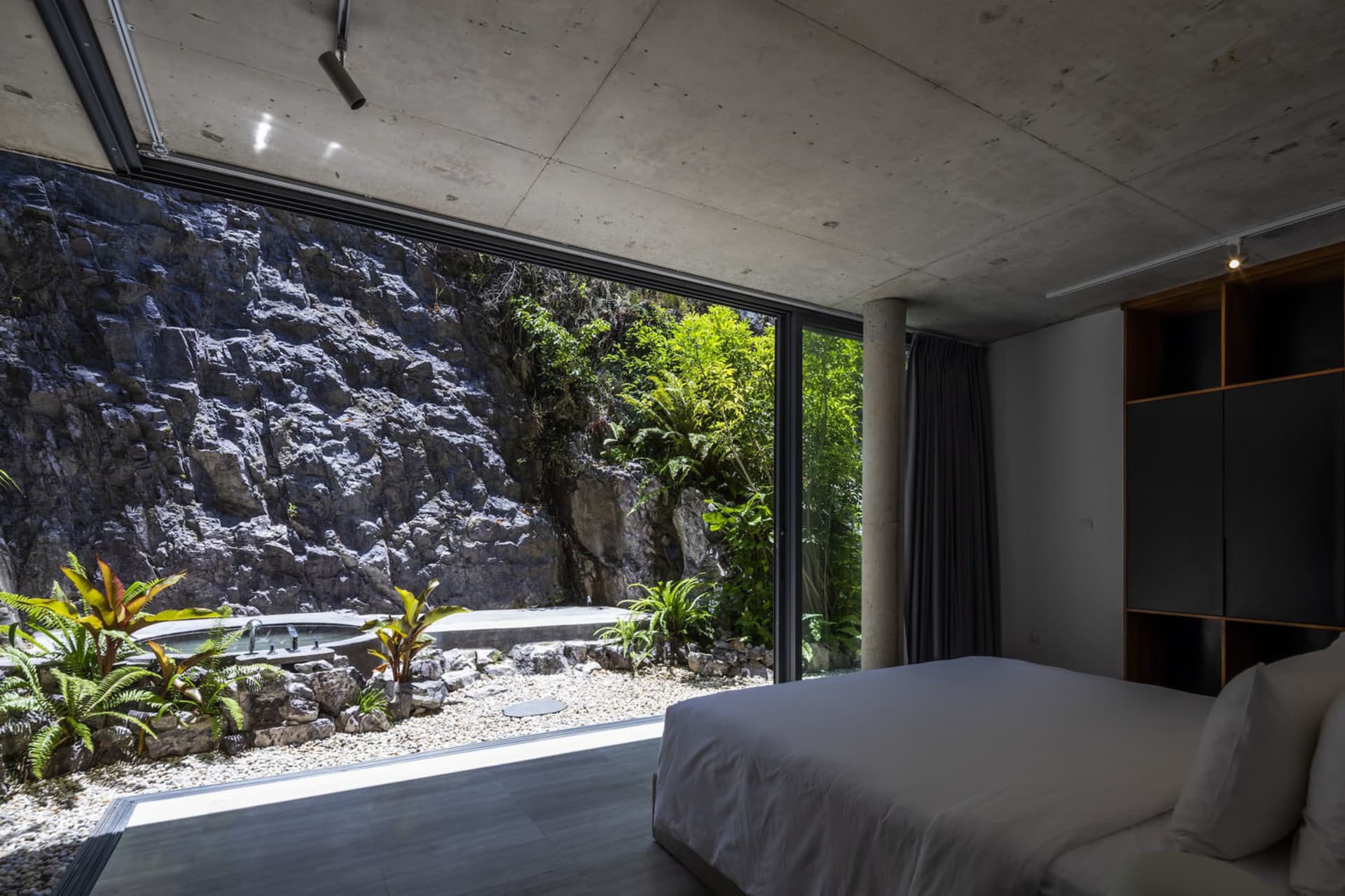 Modern industrial bedroom with concrete walls and floor, featuring a bed and large window showcasing natural surroundings.