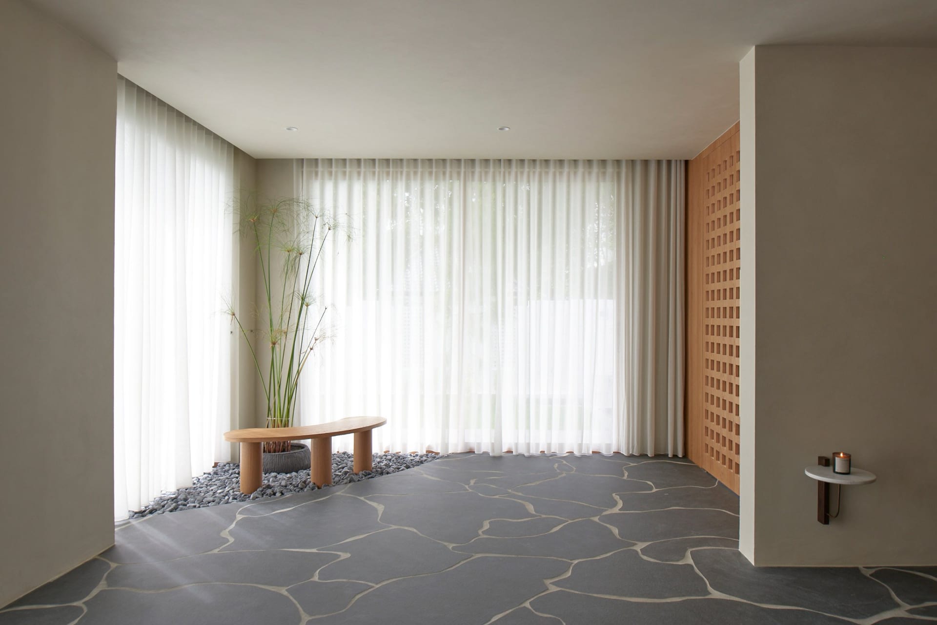 A minimalistic living room featuring a grey, stone-patterned floor, a white sheer curtain, and a wooden bench seat with a pla