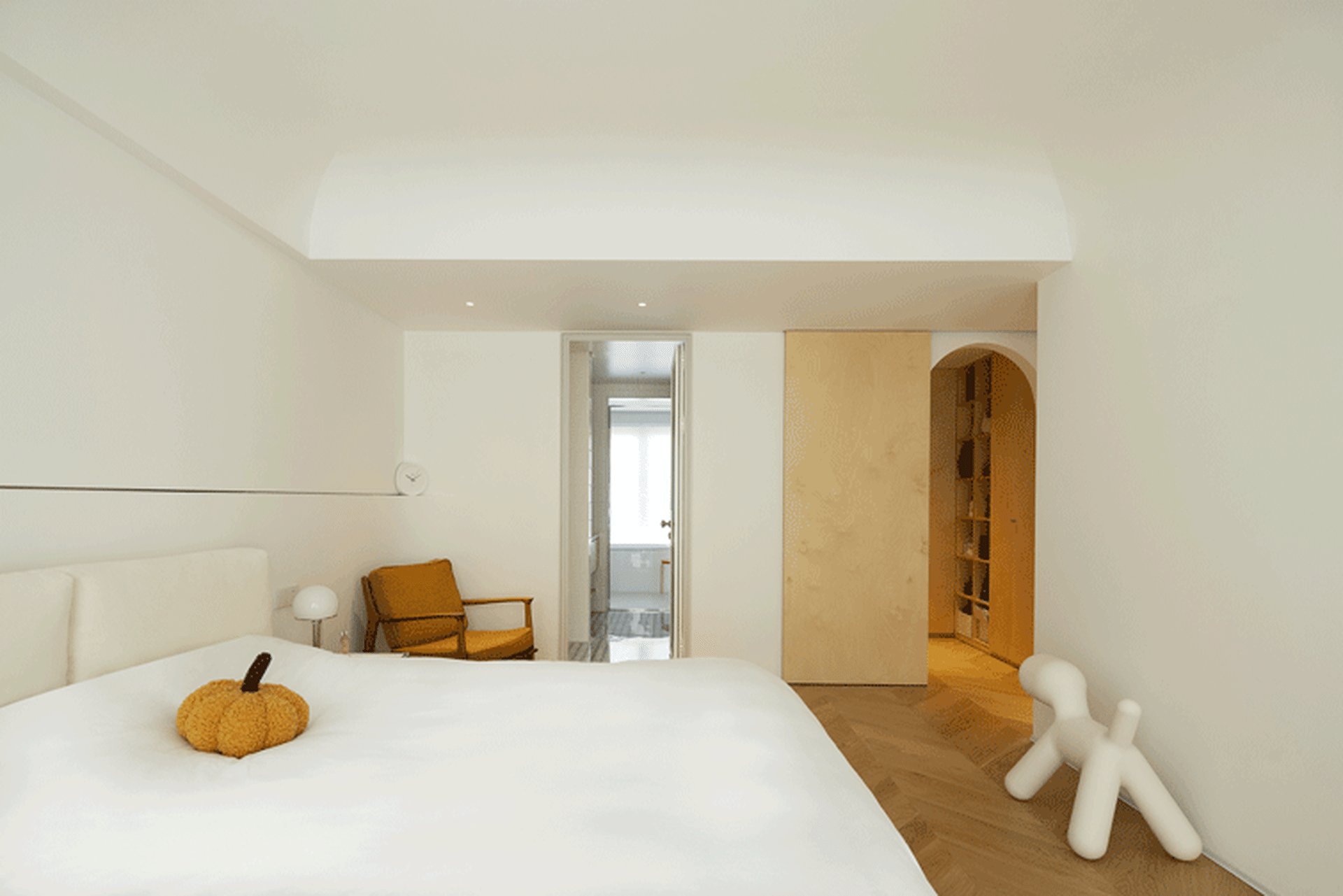 A minimalist bedroom featuring white bedding, a yellow pumpkin, and a white figure next to a wooden door.