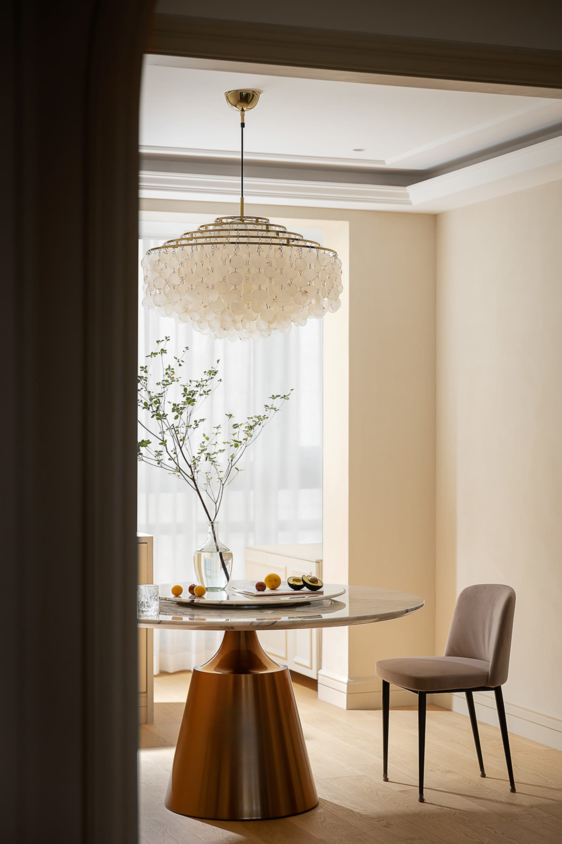 A modern residential interior featuring a round marble table, a chandelier, and a vase with green branches and fruit.