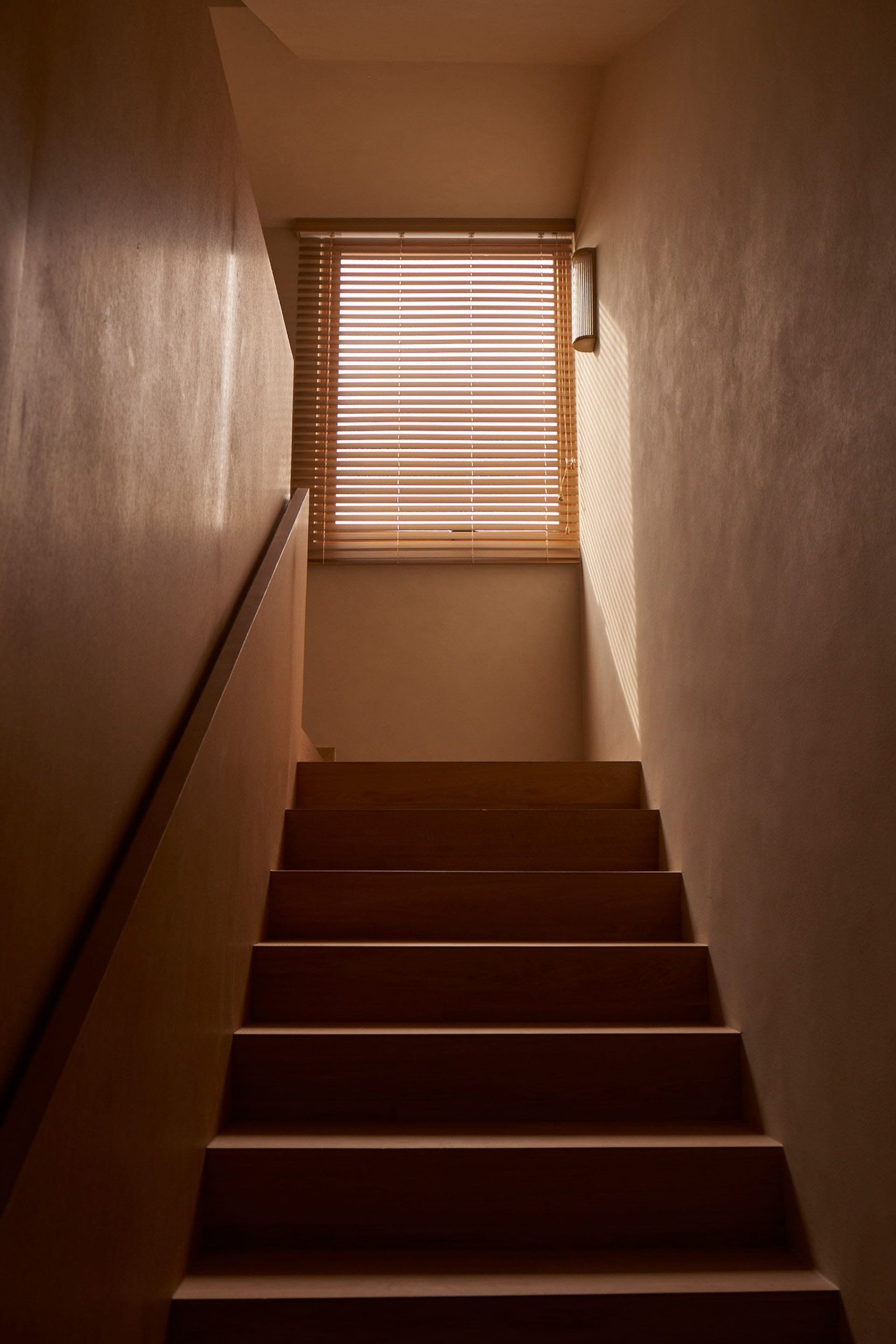 A modern staircase with wooden steps illuminated by natural light coming through a window with wooden blinds.