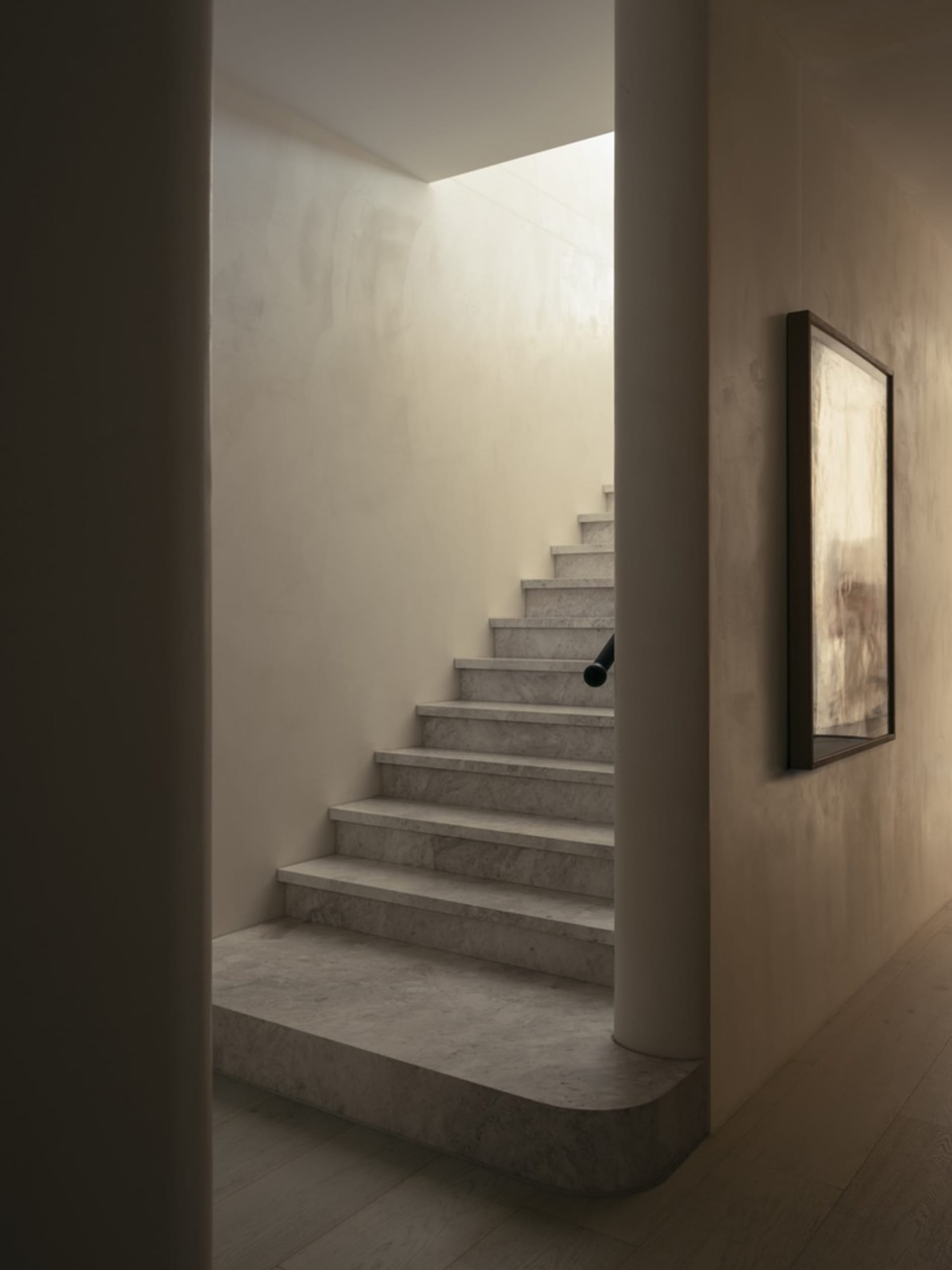 A minimalist staircase with marble steps and a white wall, illuminated by natural light from above.