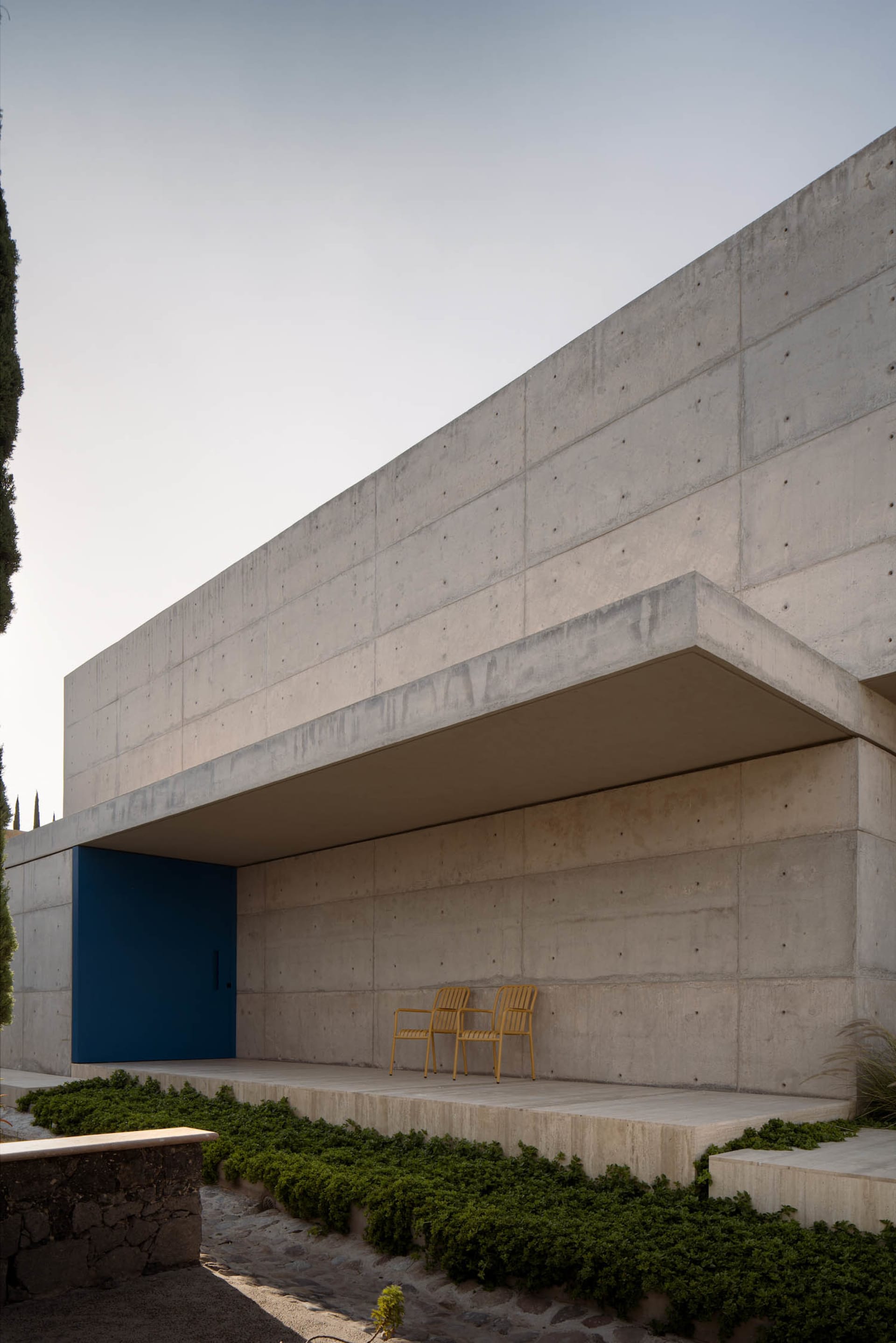 Modern building exterior with a blue door and yellow chairs on a concrete patio surrounded by greenery.