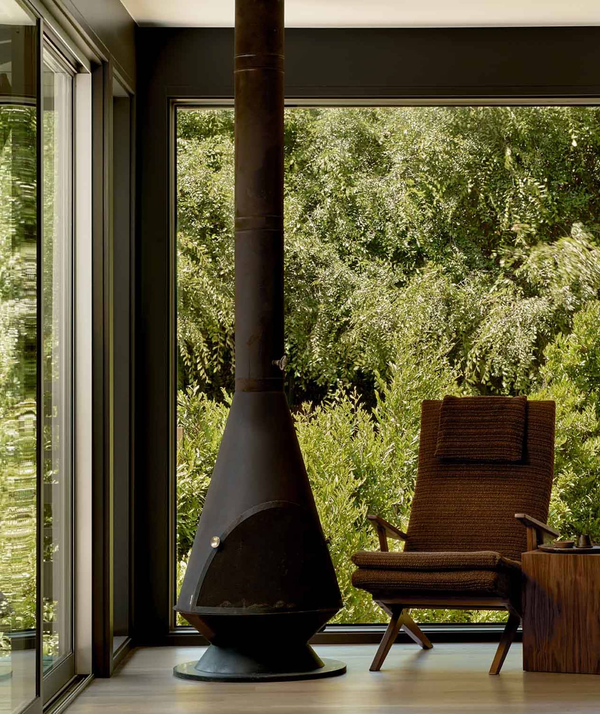 A cozy, industrial-style living room featuring a metal wood stove and a comfortable armchair surrounded by lush greenery.