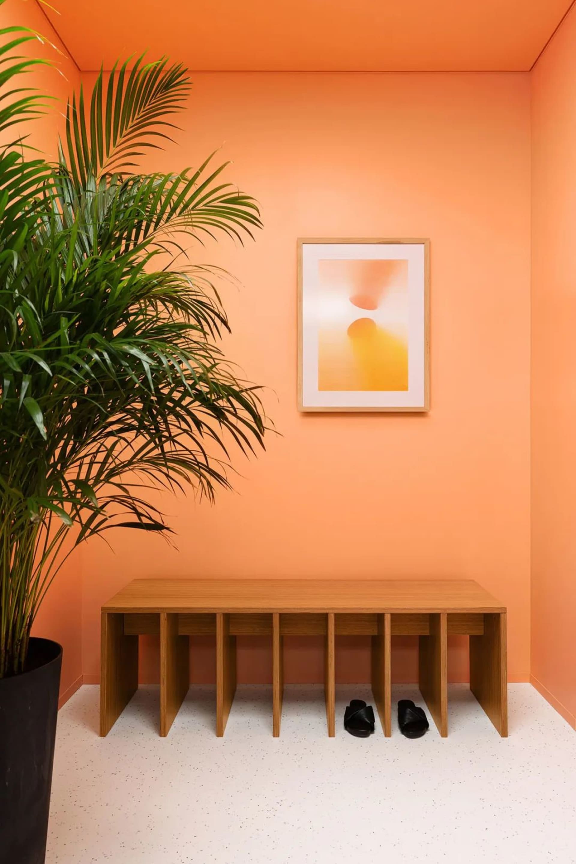 A minimalist modern entryway with a wooden shoe shelf against an orange wall, featuring two pairs of black sandals at the bas