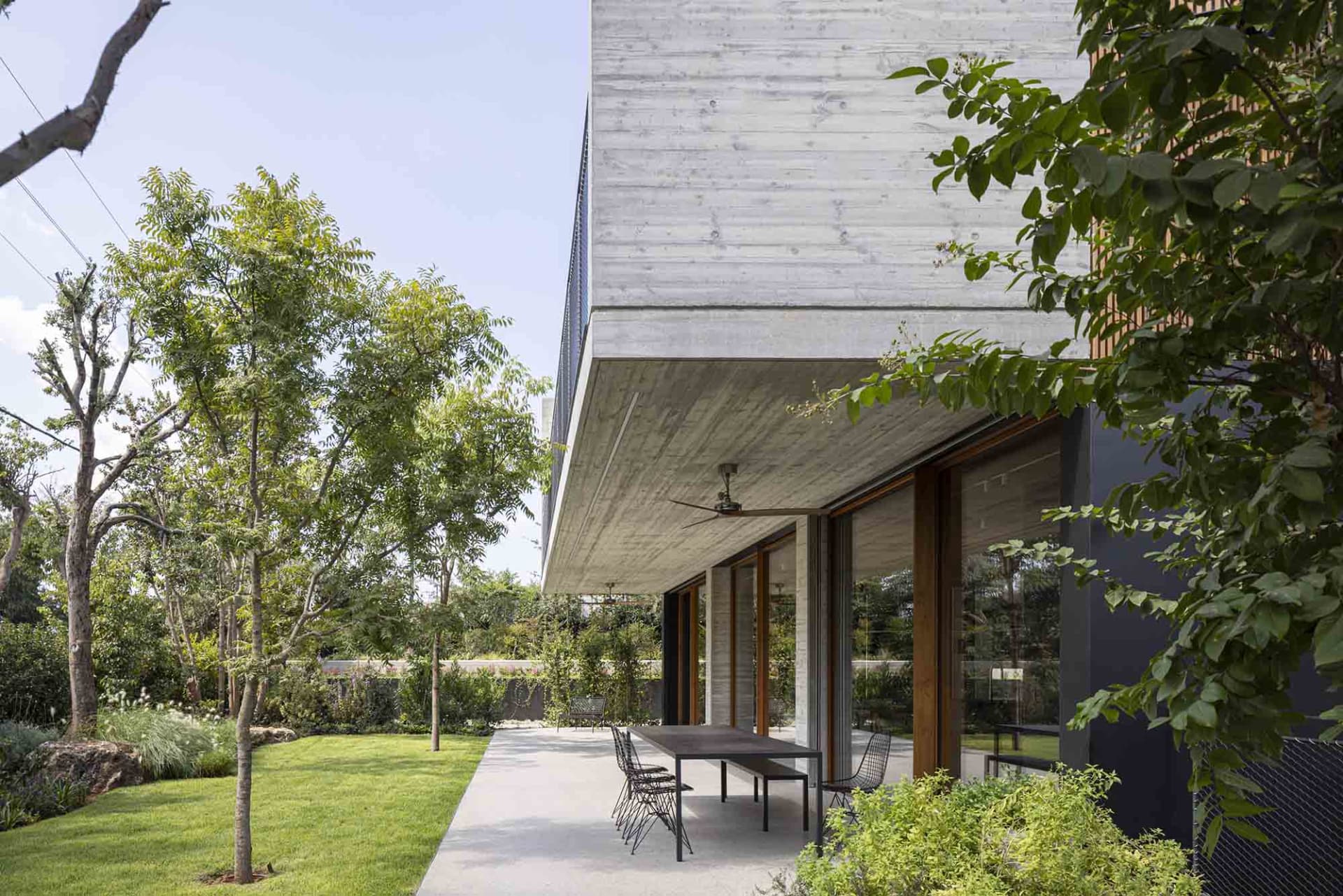 Modern house with concrete exterior featuring a patio with chairs, surrounded by greenery and trees.