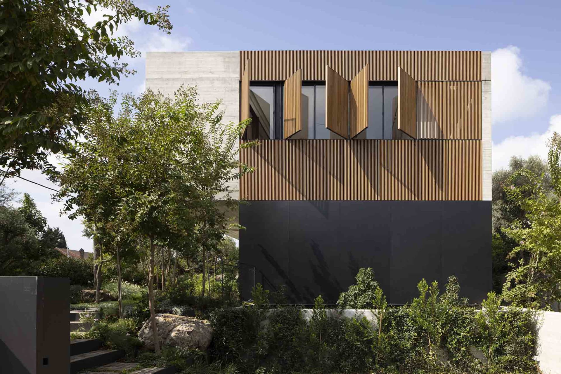 A modern house with wooden panels and concrete structure, featuring large windows with open shutters and surrounded by greene