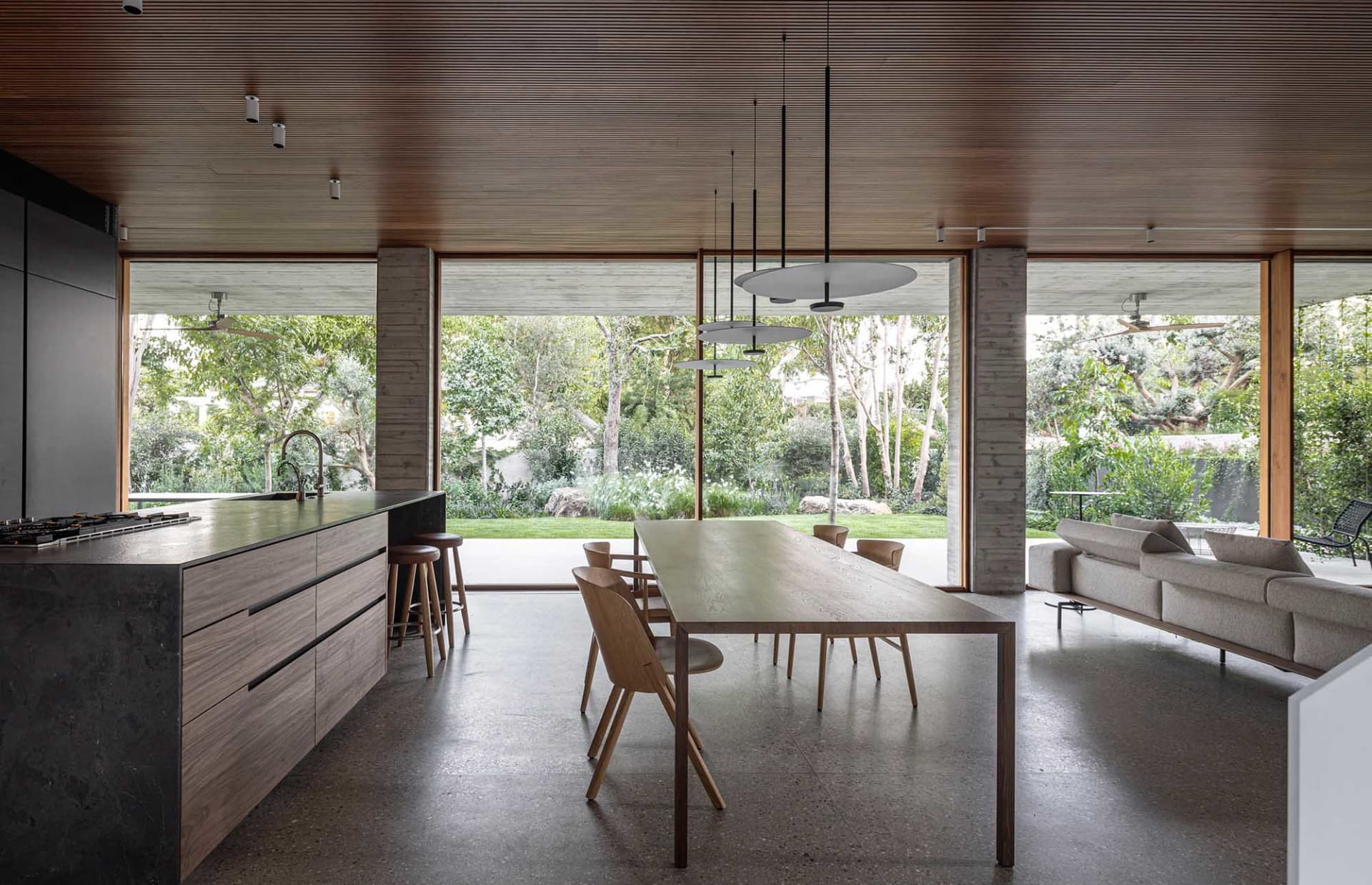 A modern residential kitchen with a wooden dining table, pendant lights, and a large window overlooking a garden.