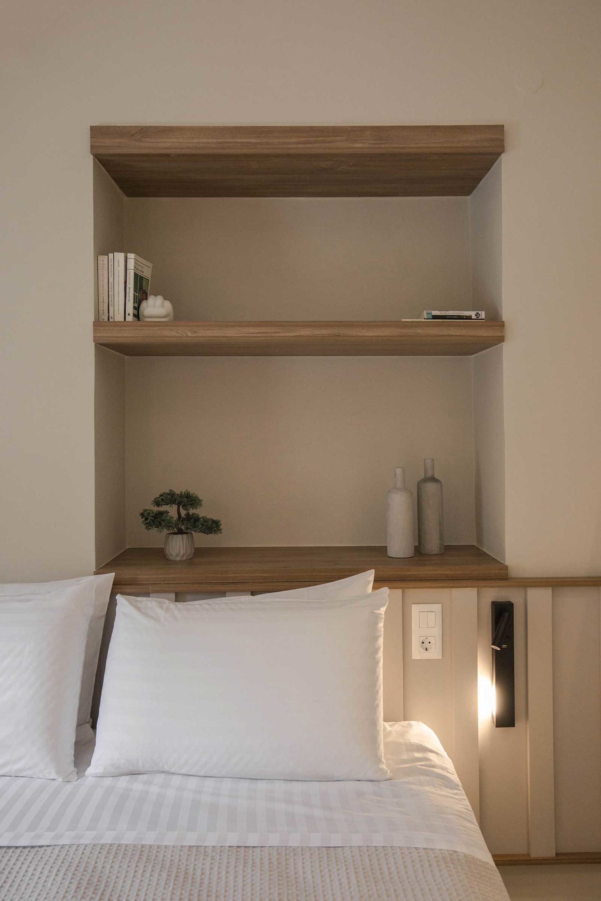 A minimalist bedroom featuring a bed with white bedding, a wooden headboard, and concrete shelves with books and decorative i