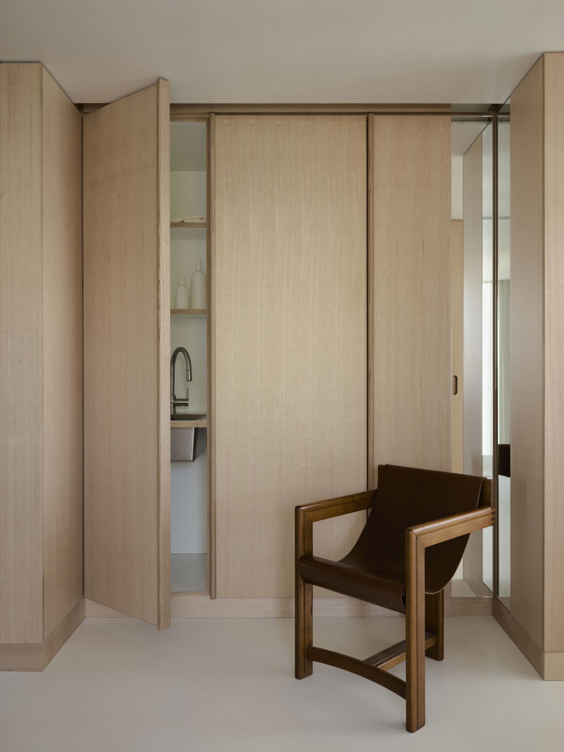 A minimalist bedroom with wooden walls and floor, featuring a chair in the foreground and a small kitchen area in the backgro