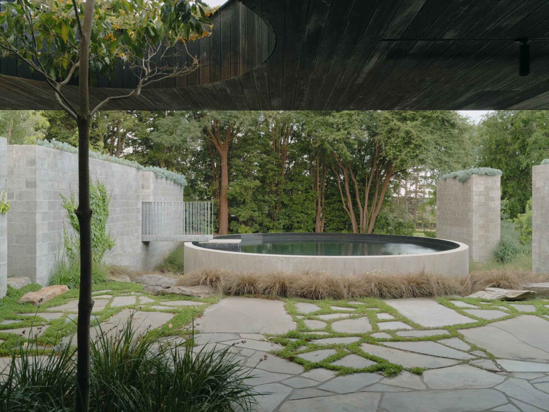 A serene outdoor space featuring a round pool surrounded by stone walls, surrounded by lush greenery and a pathway of irregul