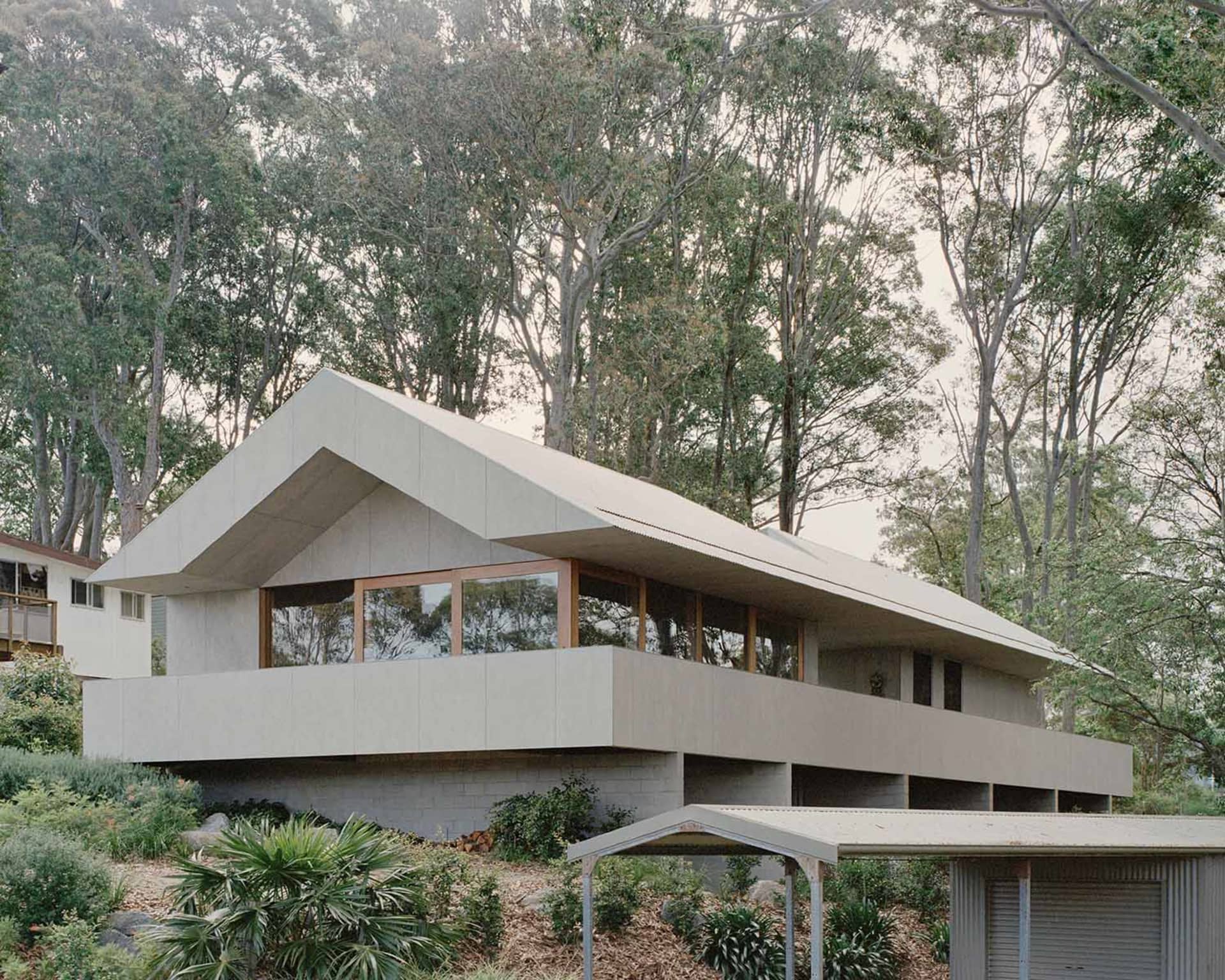 Modern concrete and metal house exterior with large windows and greenery surrounding.