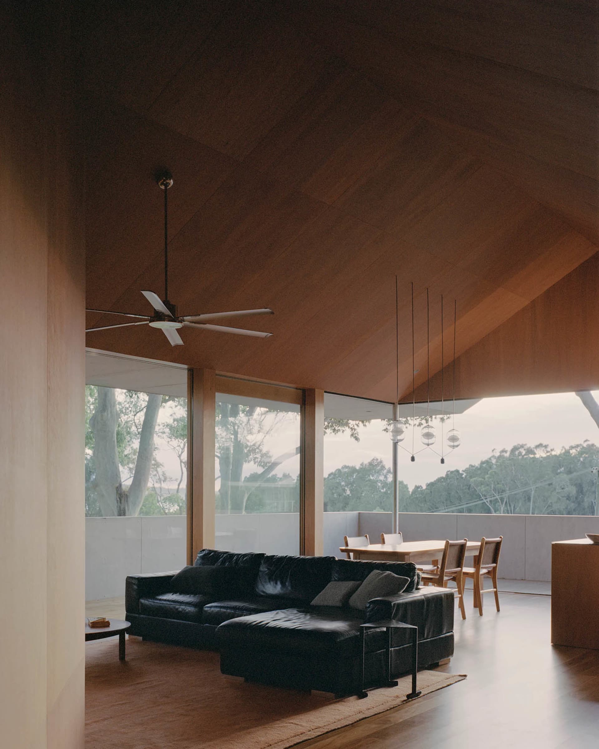 A modern living room with a black leather sofa, wooden ceiling, and large windows overlooking trees.