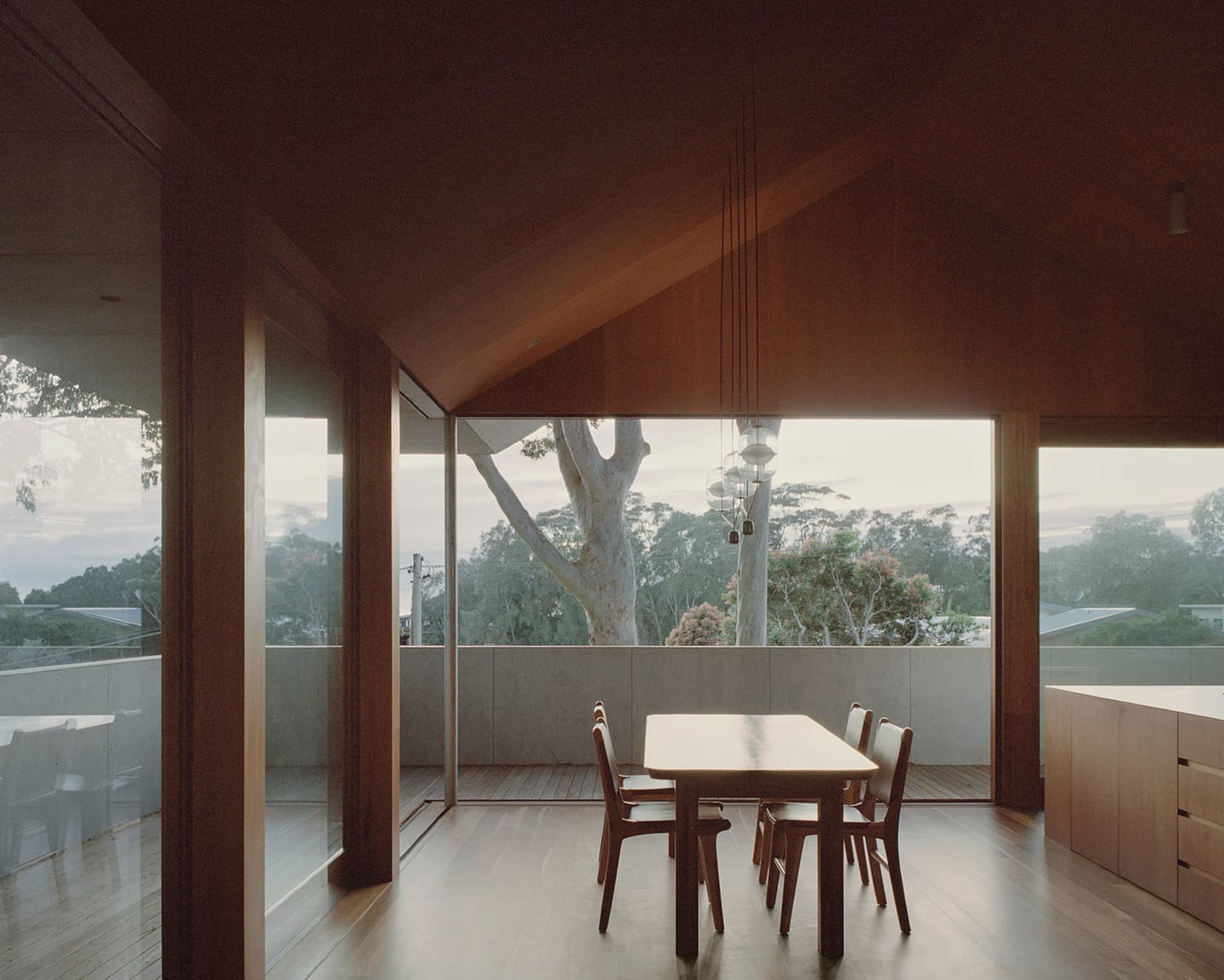 A modern residential interior with a wooden dining table and chairs, large windows, and a sloped wooden ceiling.
