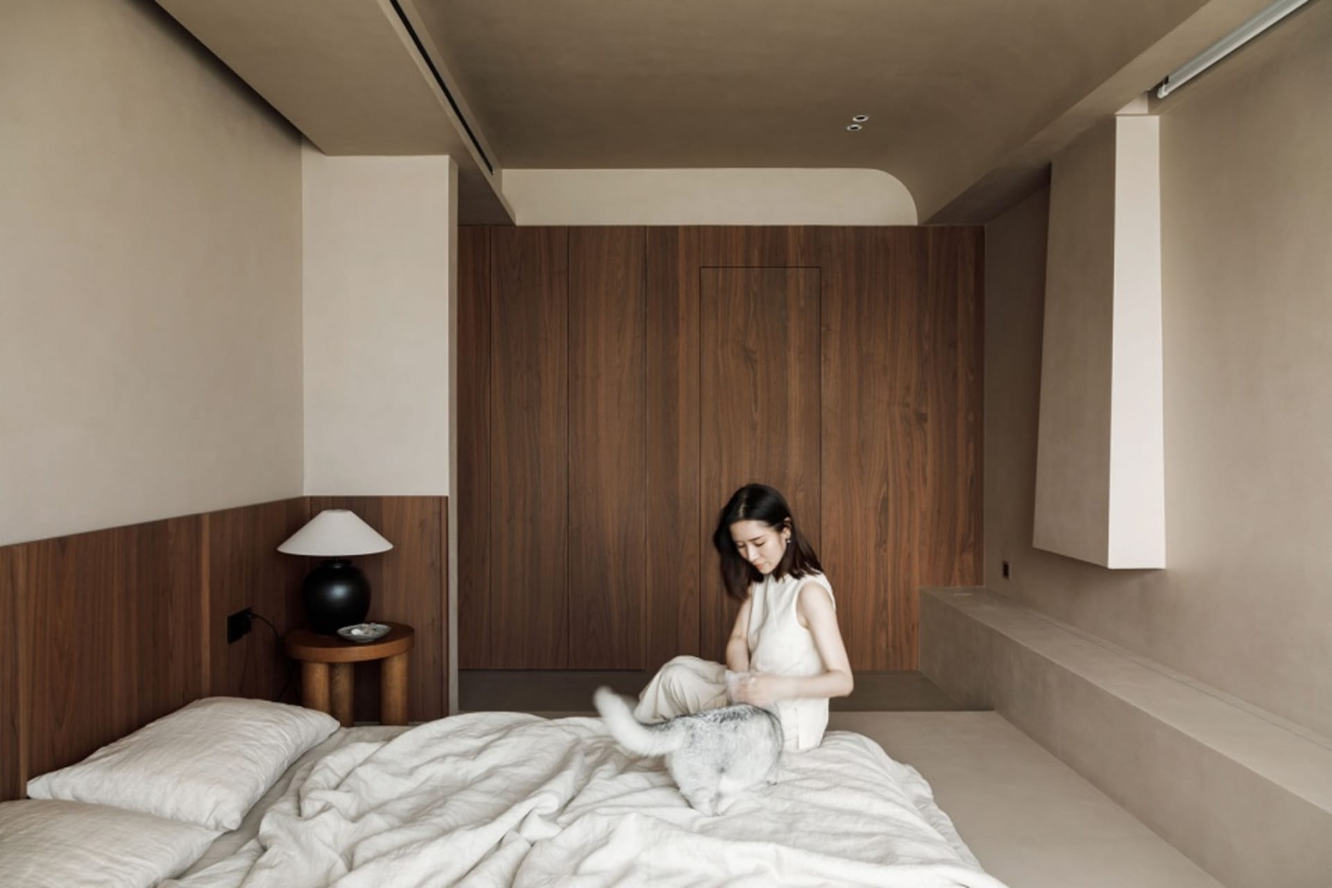 A woman sitting on a bed, petting a cat, with a bedside lamp.