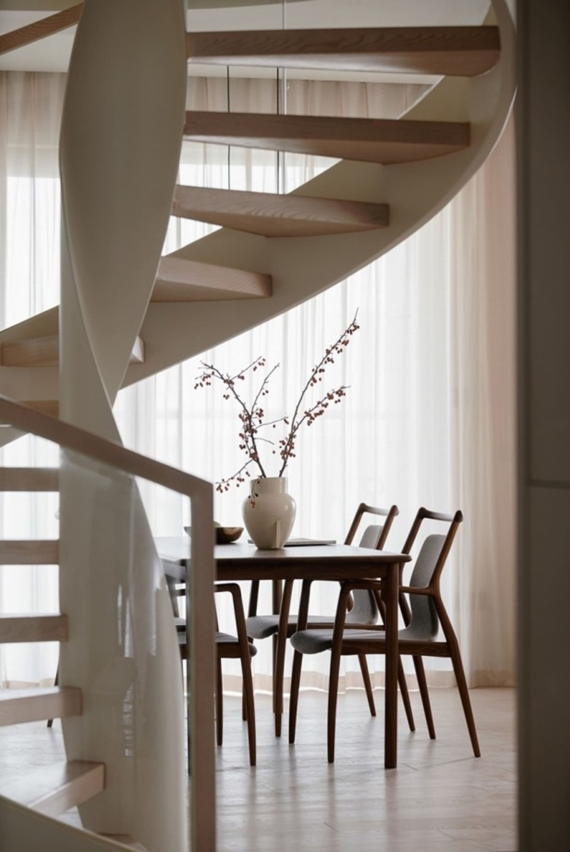 A modern minimalist dining room featuring a wooden table, chairs, and a vase with branches.