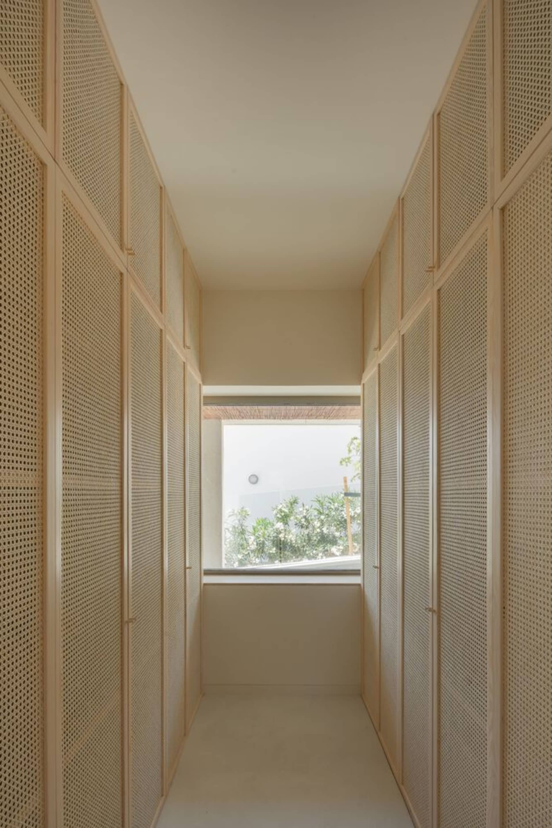 A narrow, light wood corridor with transparent partitions leading to a window view of a green outdoor space.