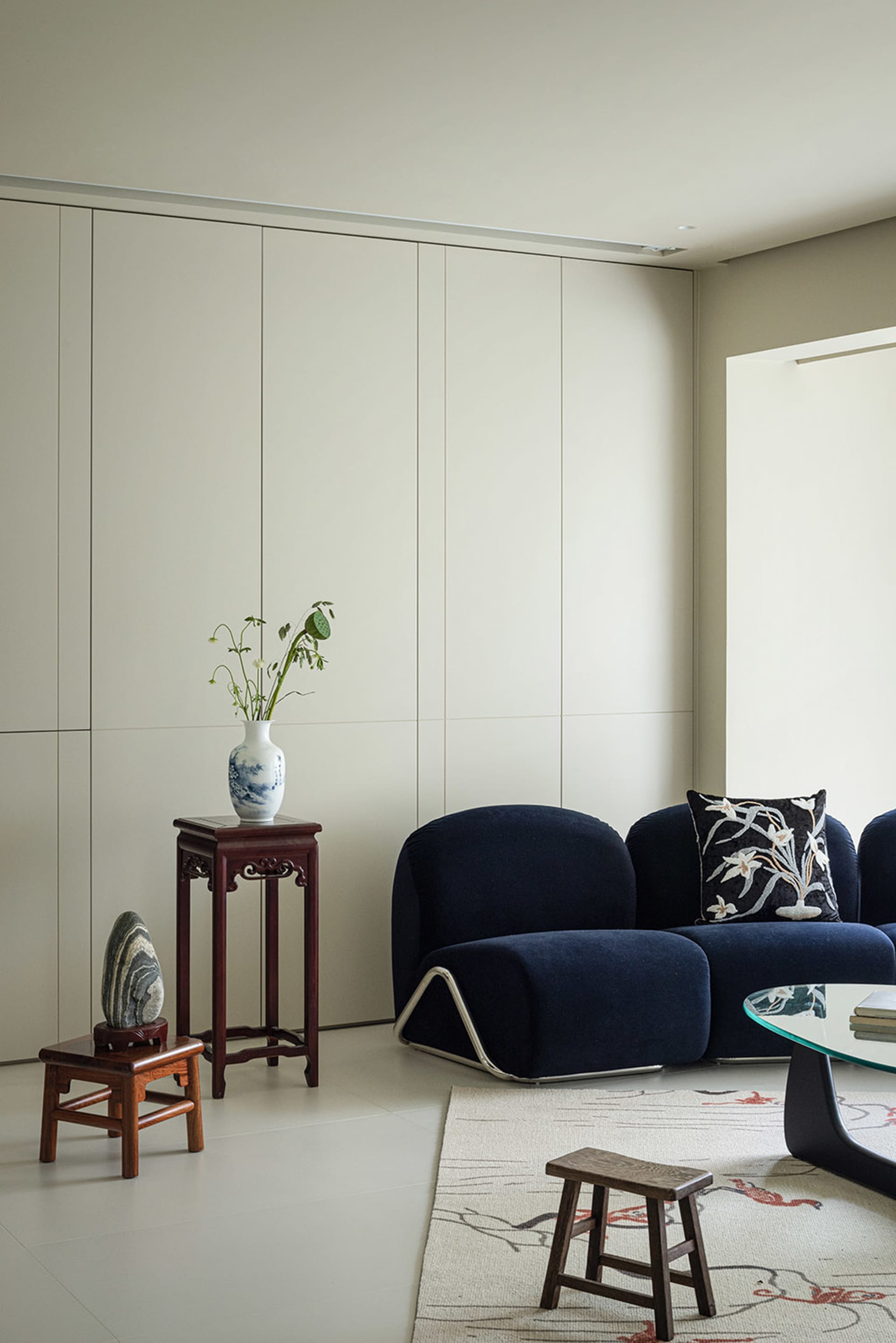 A modern living room featuring a blue fabric couch with a white pillow, a white vase with flowers on a wooden pedestal, and a