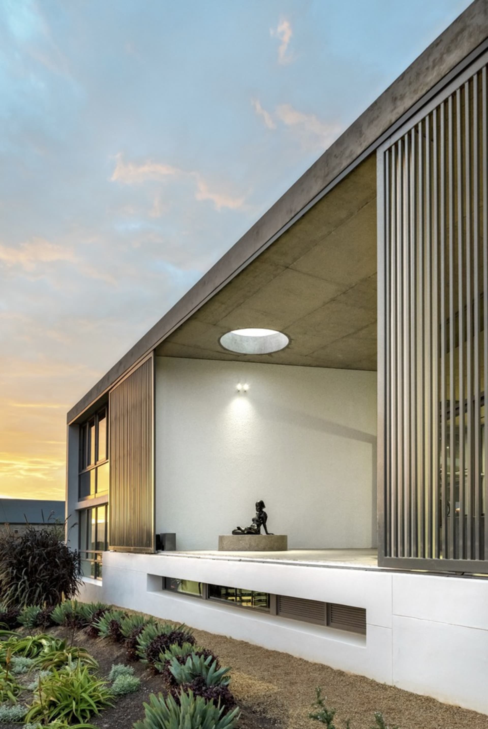 A modern building with a concrete exterior and metal accents, viewed at sunset with a circular skylight and sculpture in the