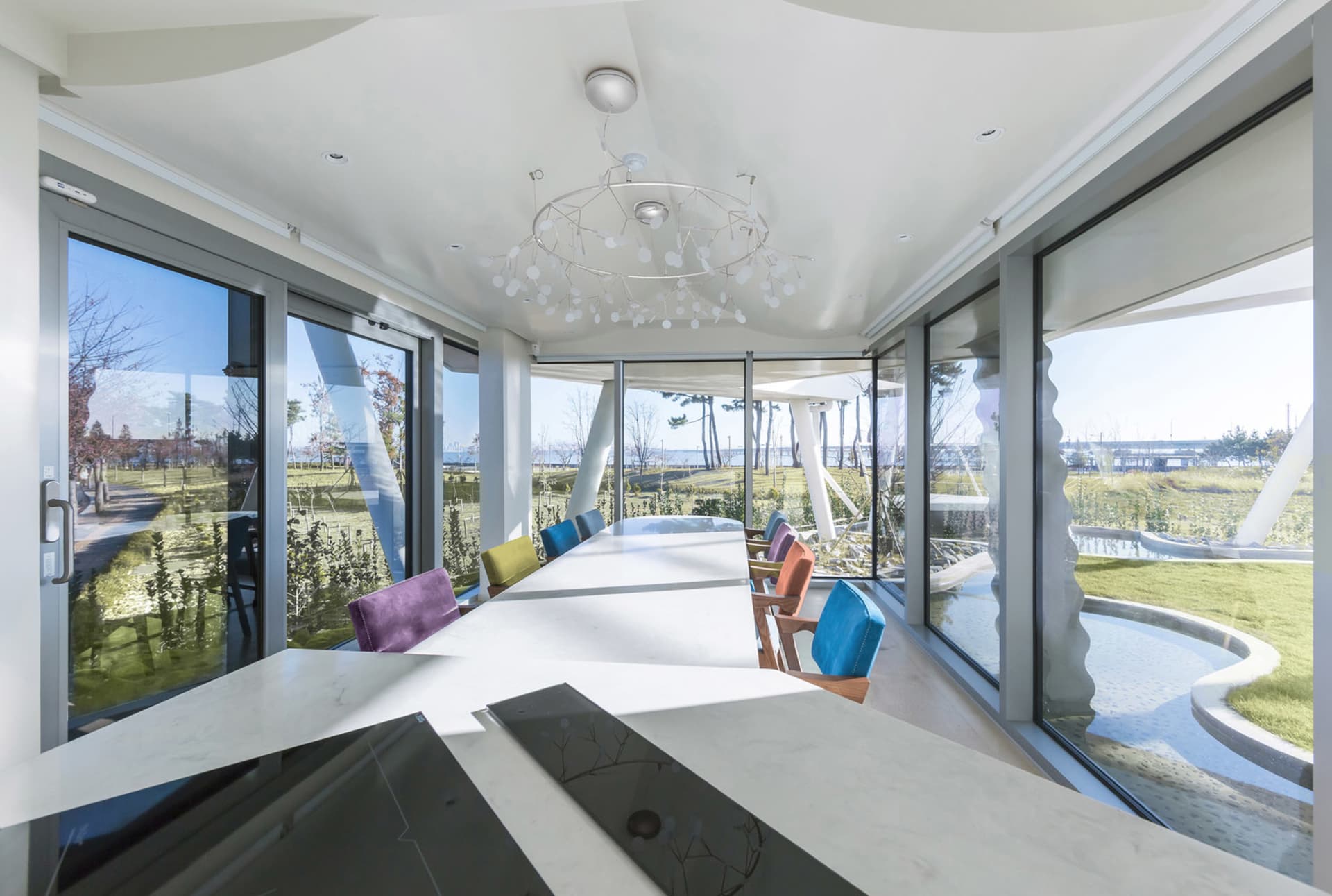 A modern dining room with a long table surrounded by colorful chairs, featuring large glass windows overlooking a green lands