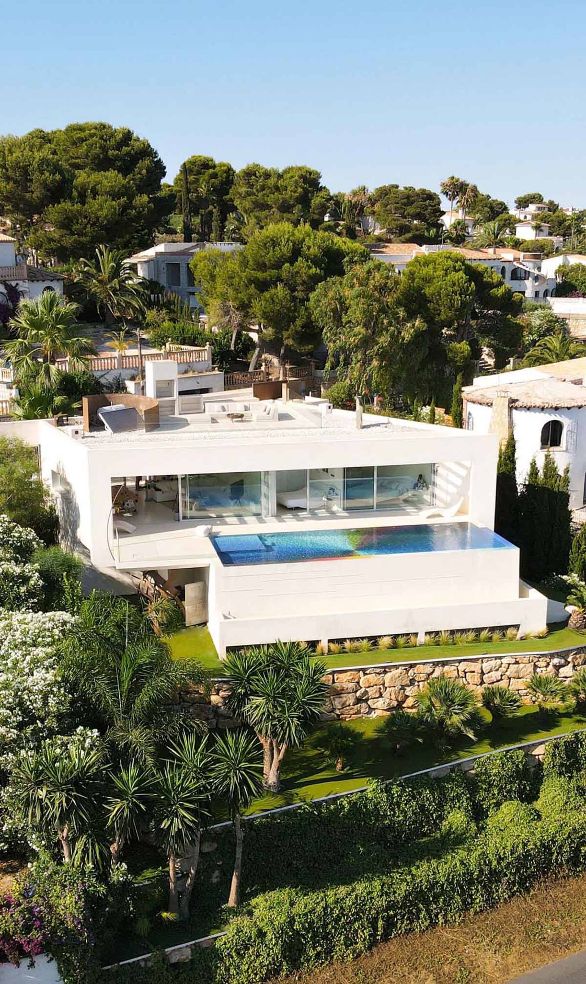 Modern white villa with glass walls and stone accents, surrounded by lush greenery.
