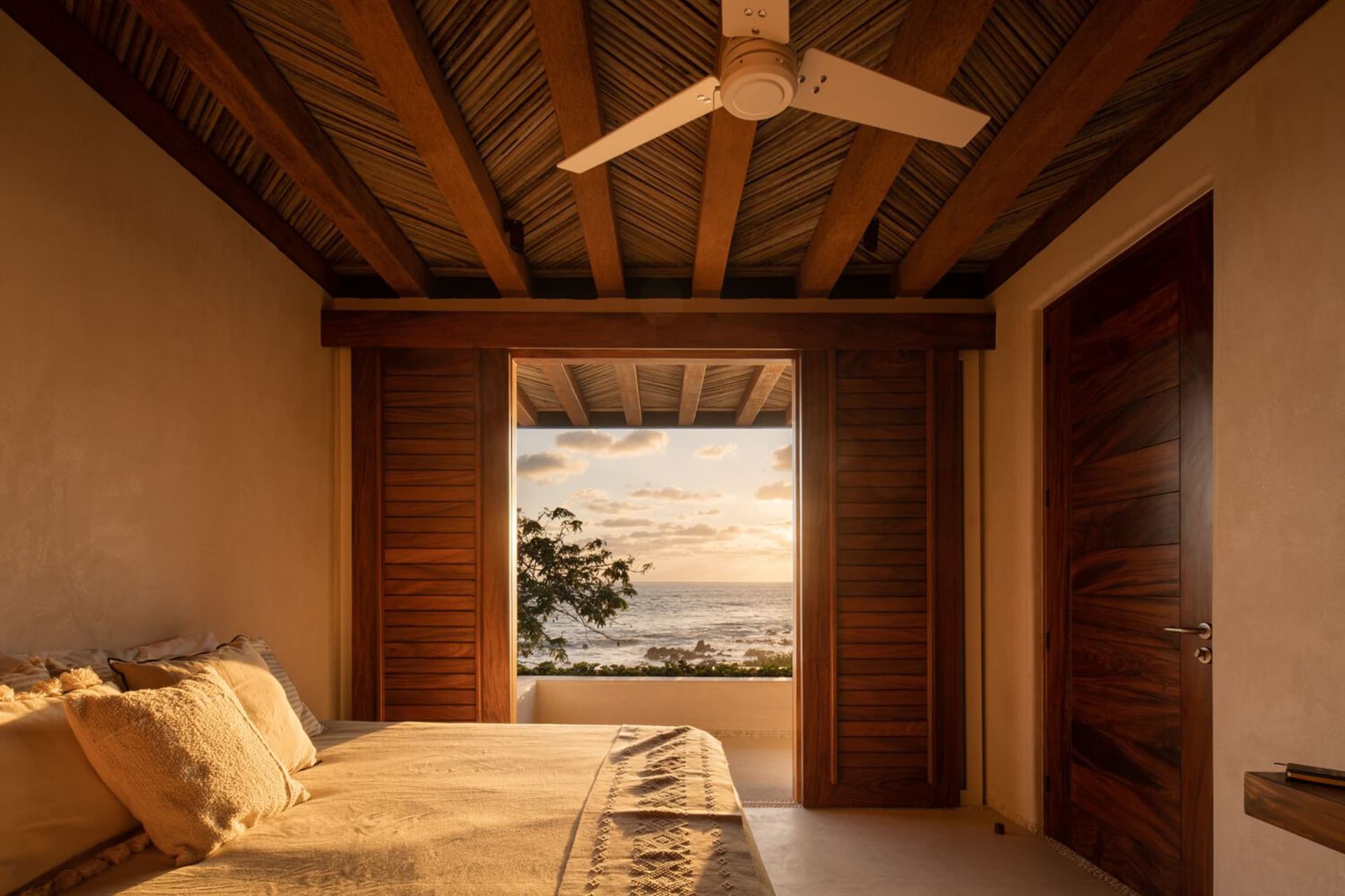 A cozy bedroom featuring wooden beams on the ceiling, a bed with fabric bedding, and a coastal view through the window.