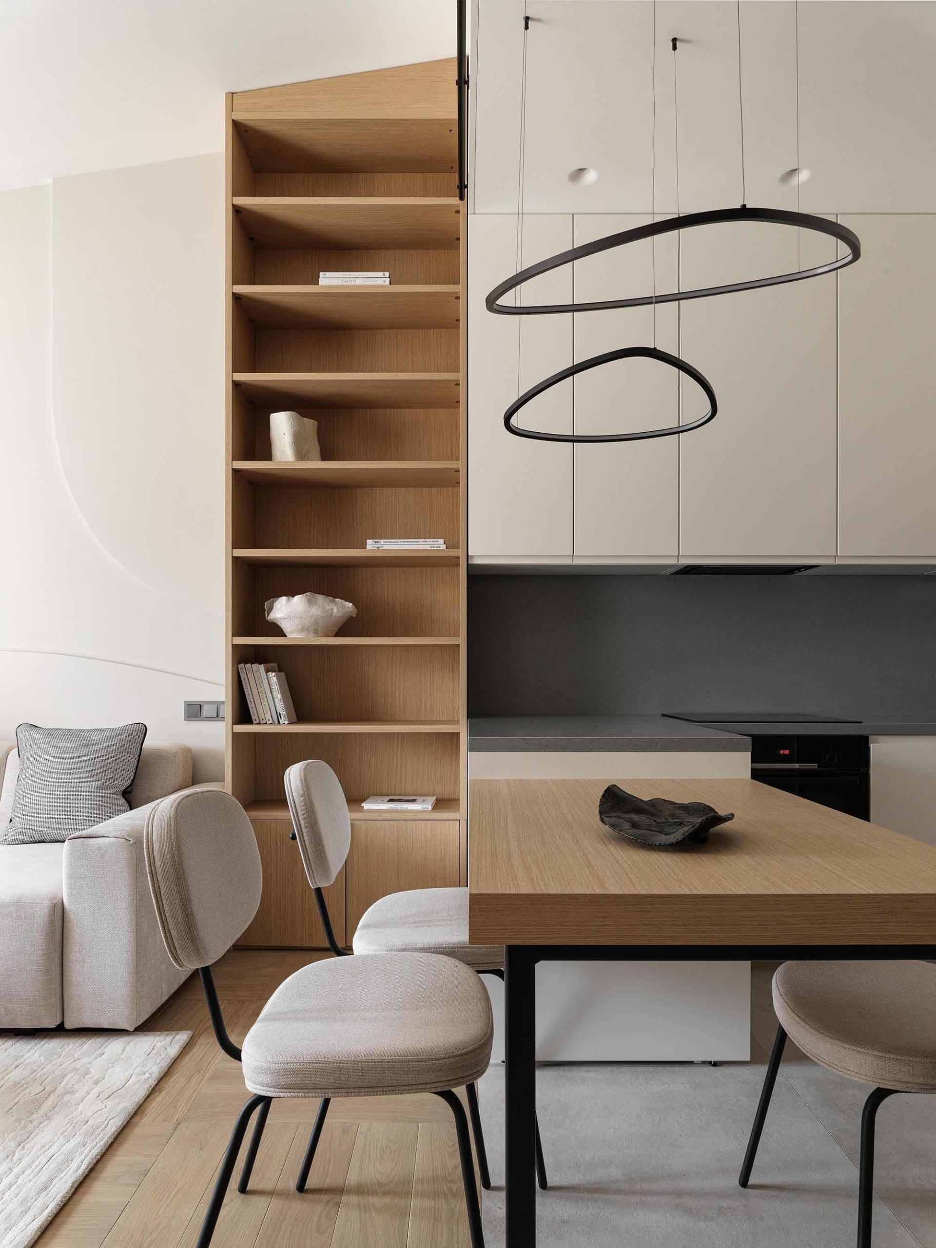 A modern minimalist kitchen featuring light-colored wooden shelves and a dining table with contemporary lighting fixtures.
