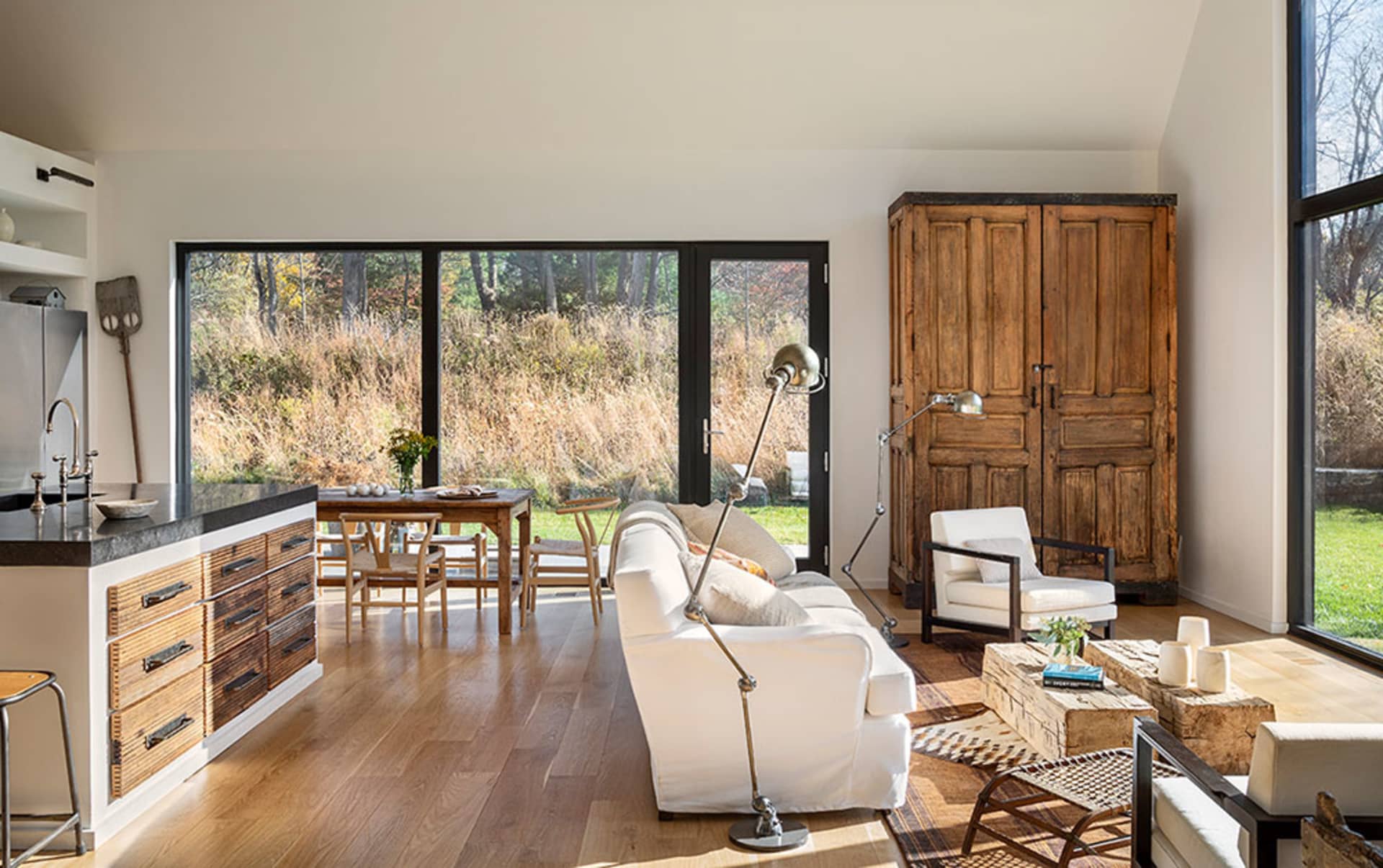 A modern living room with a white sofa, wooden floor, and large windows showcasing a natural landscape.