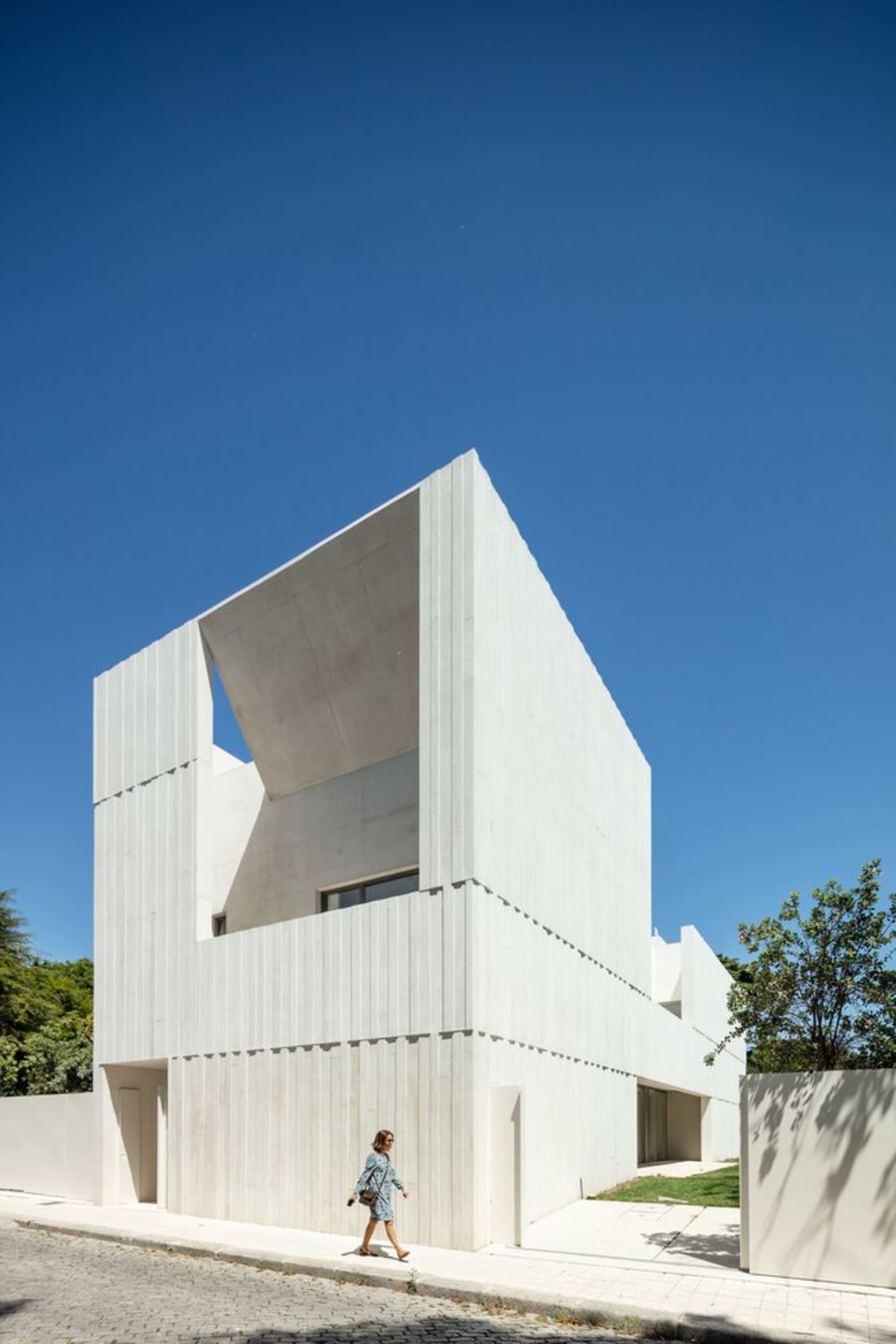 A modern white concrete building with an open space on the top floor and trees in the background.